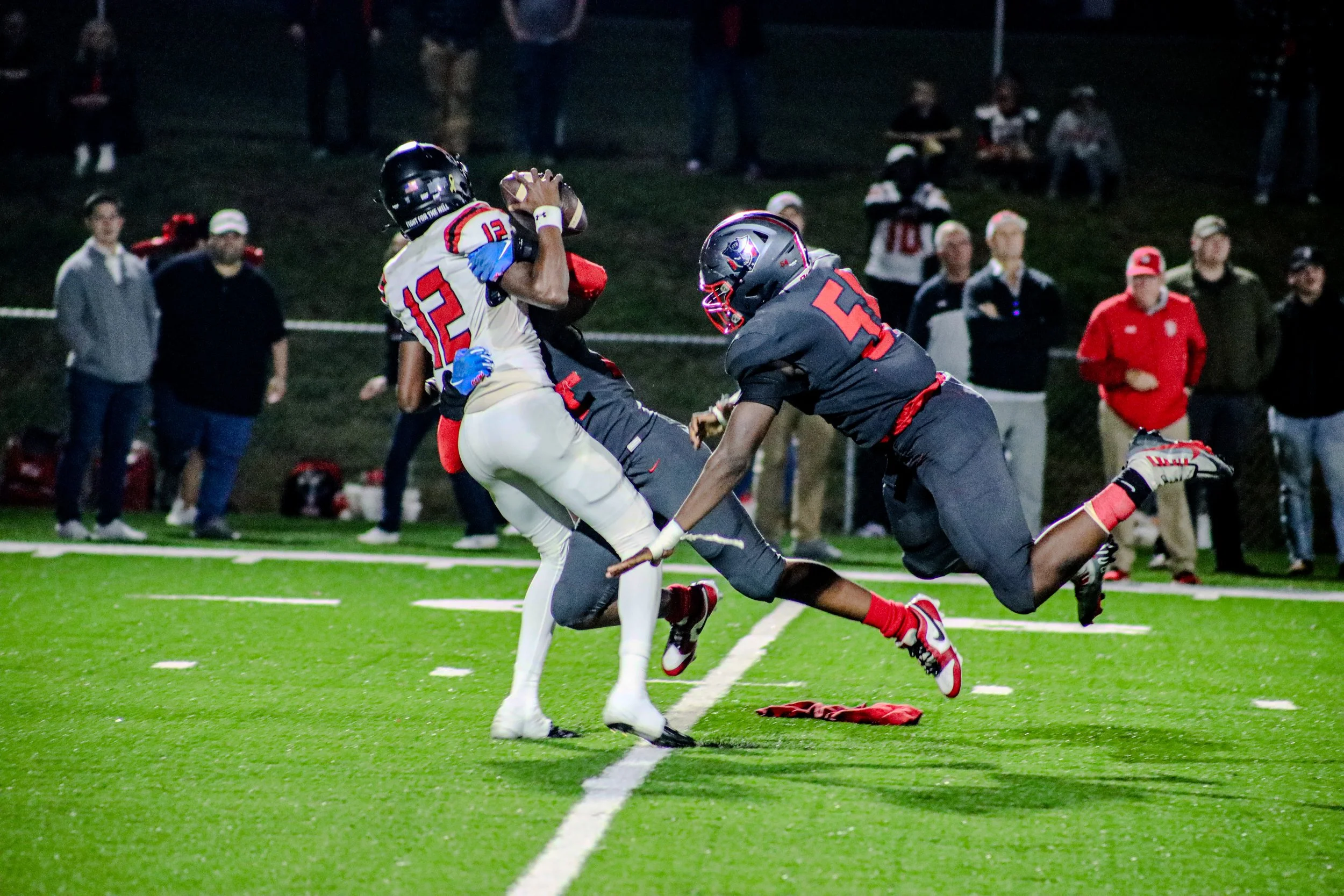 High school football player in white jersey catches the ball as a defender in navy attempts a tackle under stadium lights