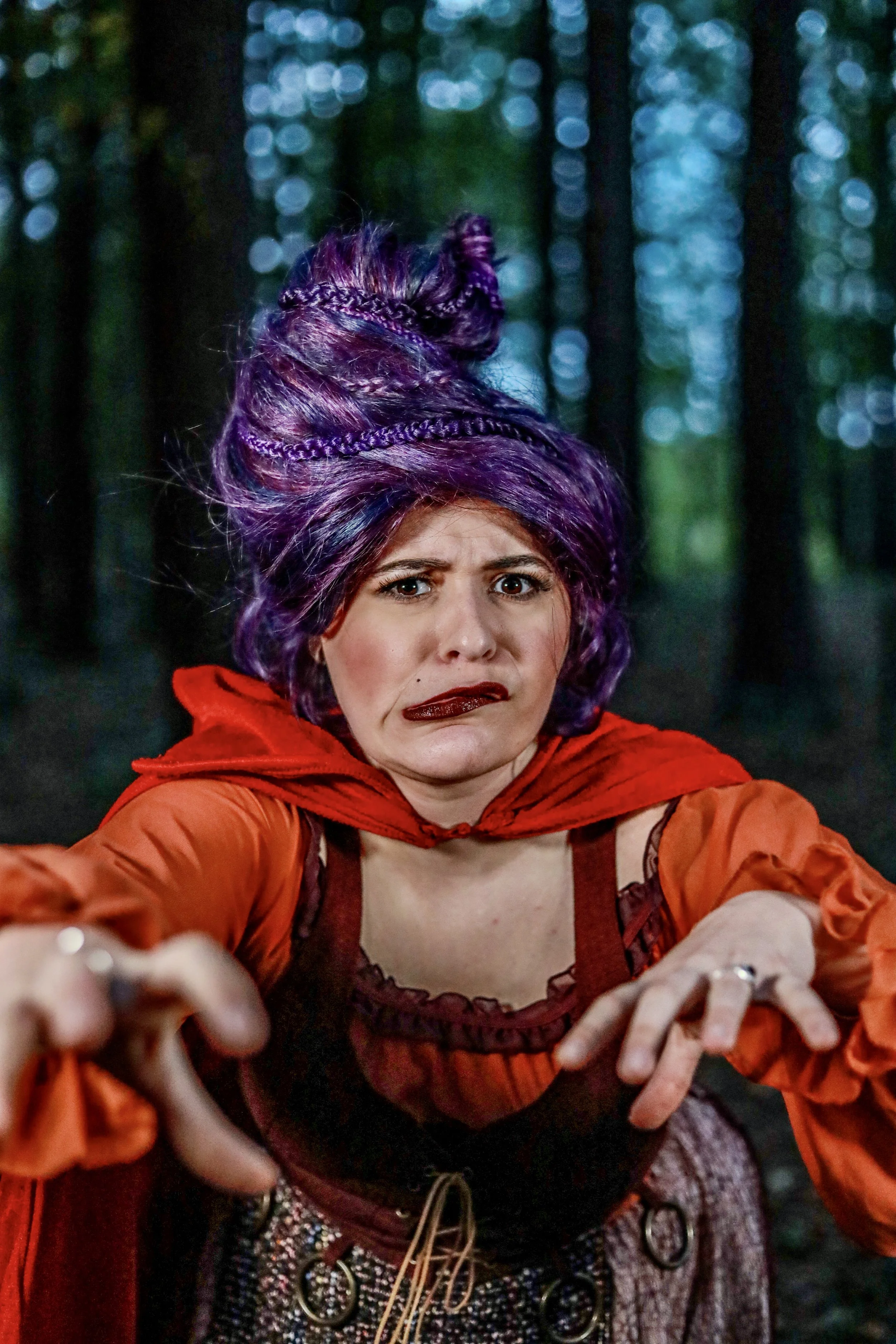 Woman with purple braided hair wearing a red and orange costume against a blurred forest background