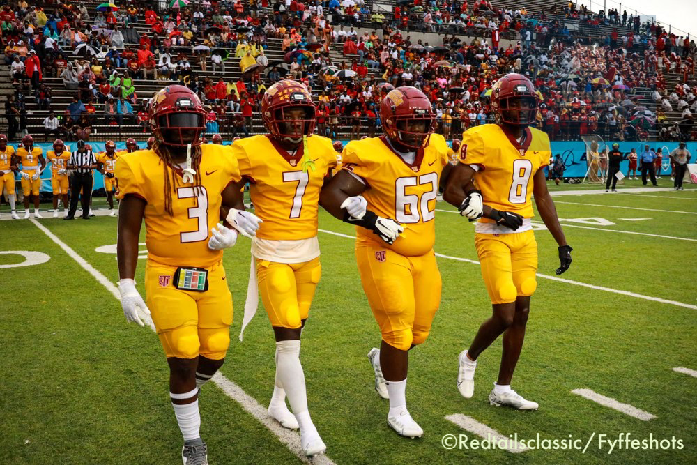 Four college football players in yellow uniforms and helmets walk across the field as a packed crowd watches from the stadium stands.
