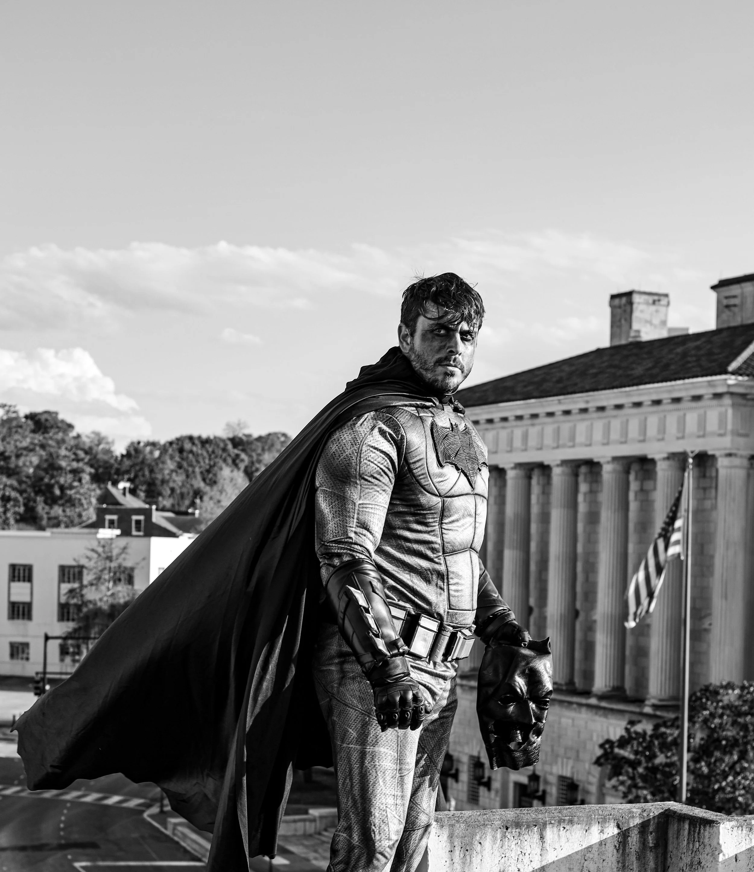 Black-and-white photo of a costumed superhero in a cape and armored suit standing on a rooftop with a city building behind him