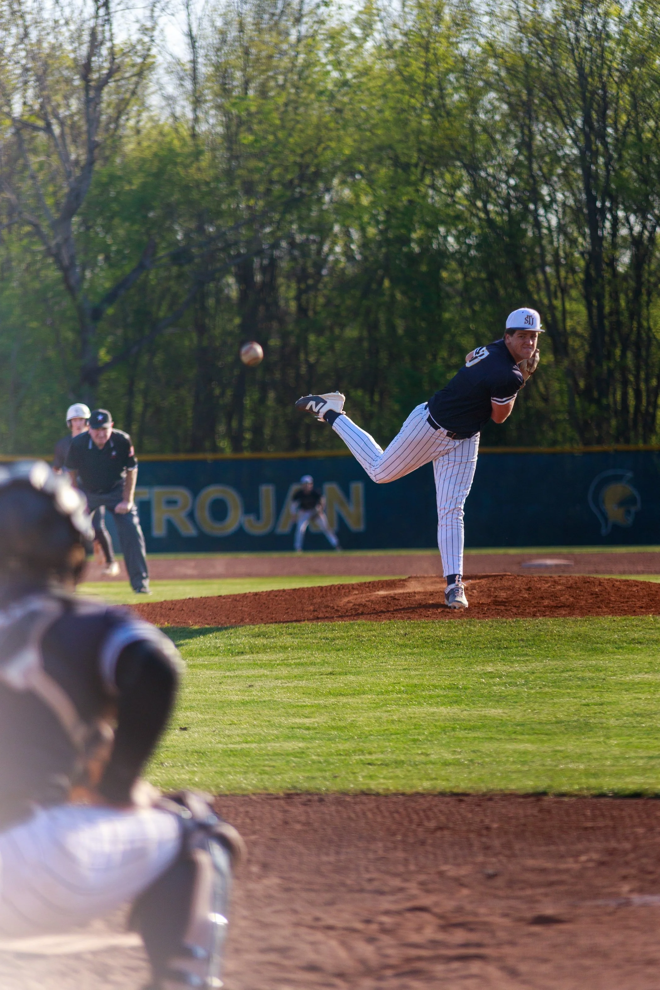 Baseball pitcher in a dark jersey and pinstriped pants delivers a pitch on an outdoor field.