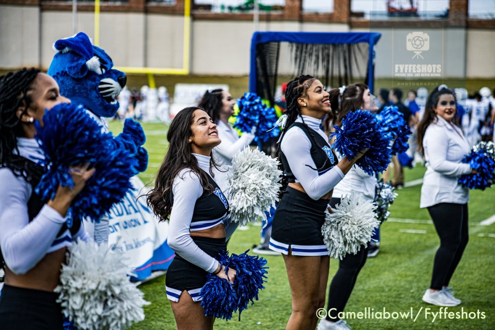College cheerleaders holding blue and white pom-poms perform on a football field alongside a blue mascot.