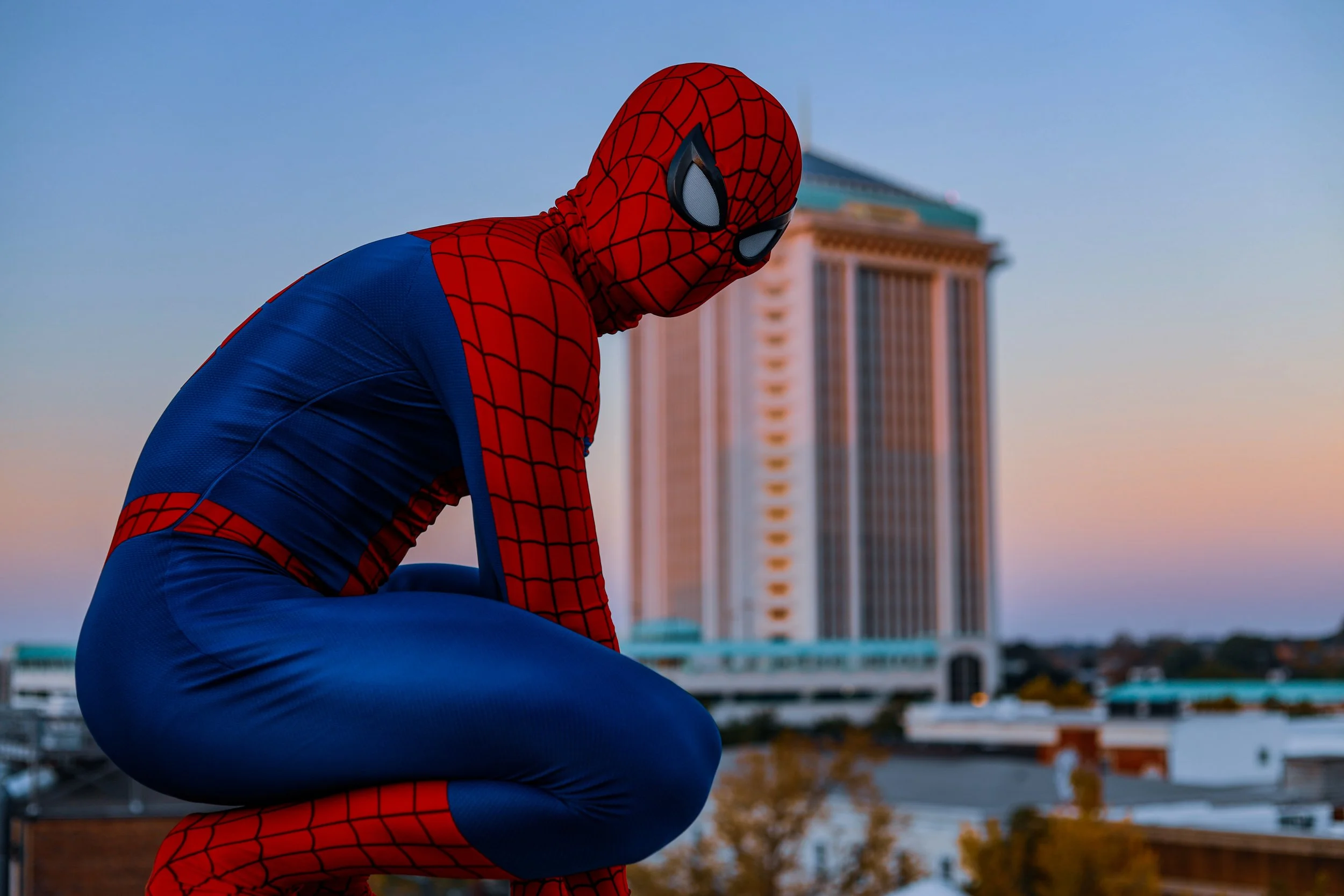 Person in a Spider-Man costume crouching on a rooftop with a city building in the background