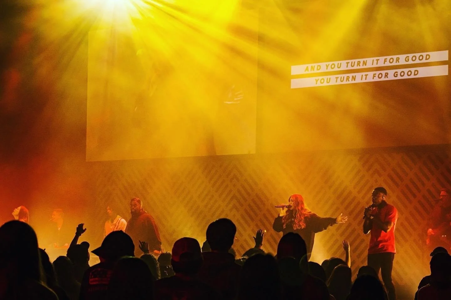 Live worship concert with singers performing under golden stage lights and audience members raising their hands