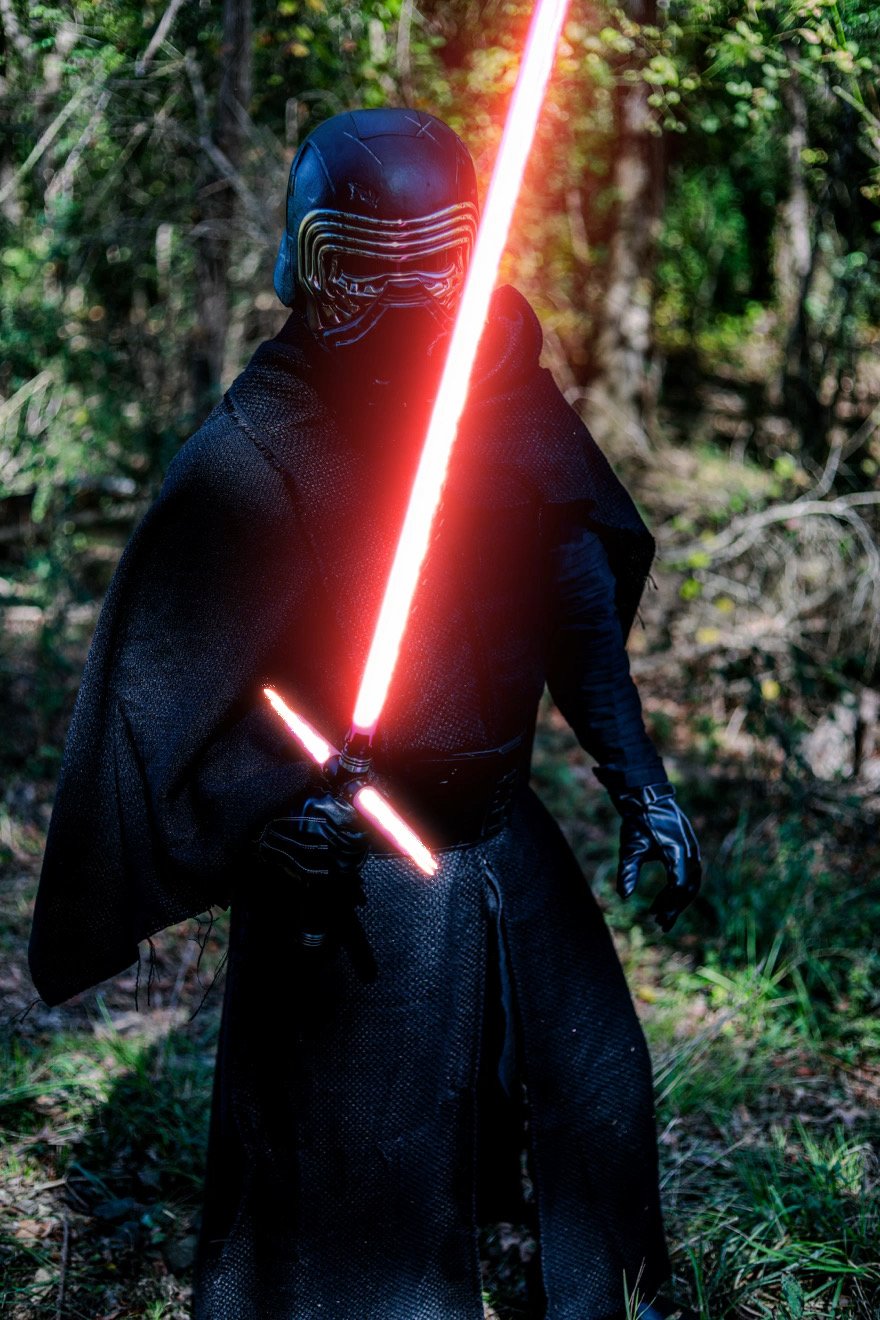Person dressed as Kylo Ren holding a glowing red crossguard lightsaber in a forest