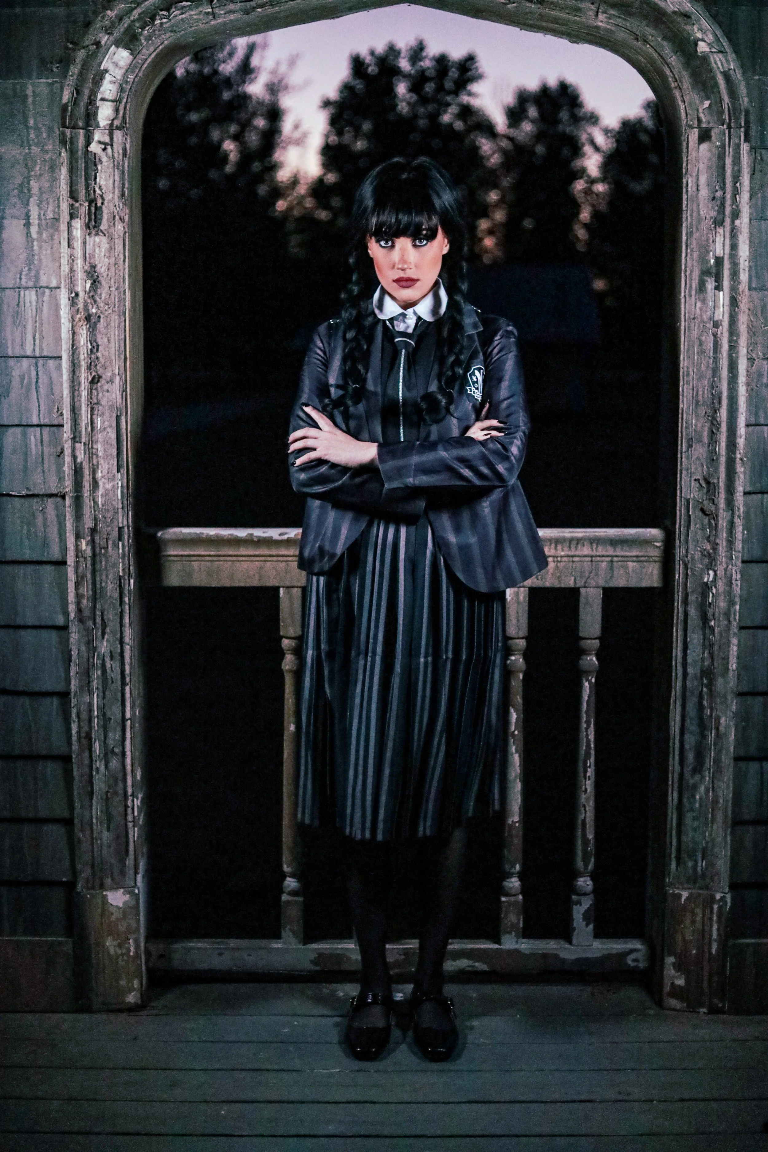 Gothic woman with braided black hair and arms crossed, standing in an arched window wearing a black blazer and striped skirt.