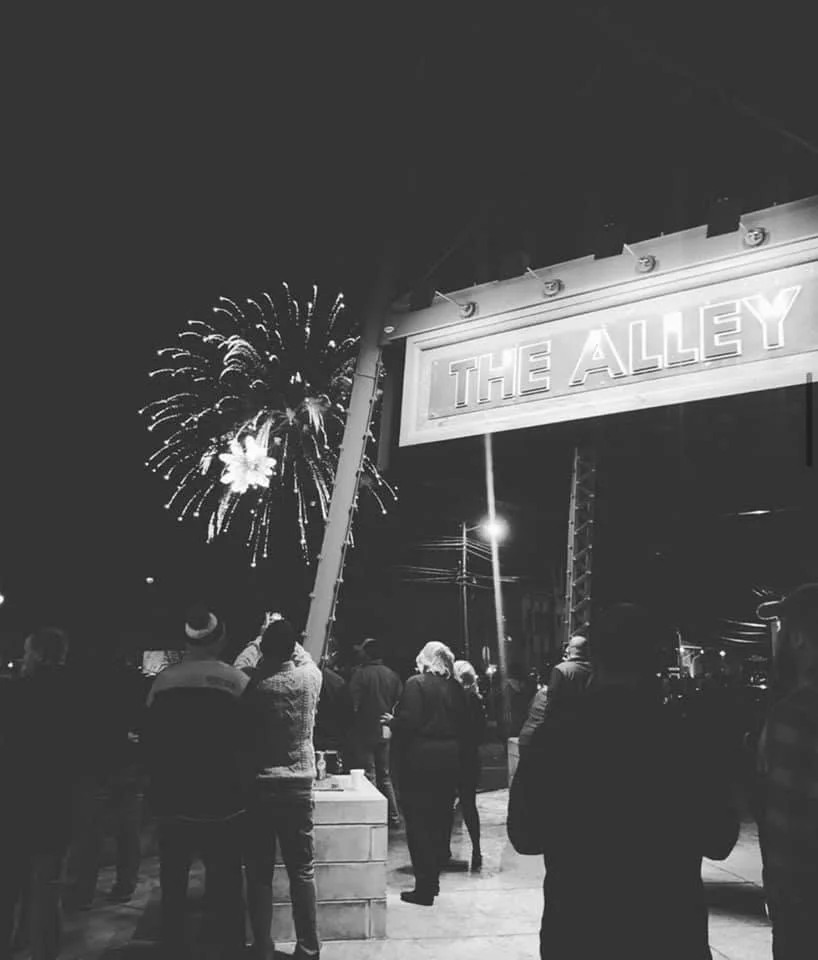 Black-and-white night scene of fireworks above a crowd beneath a lit "The Alley" entrance sign.