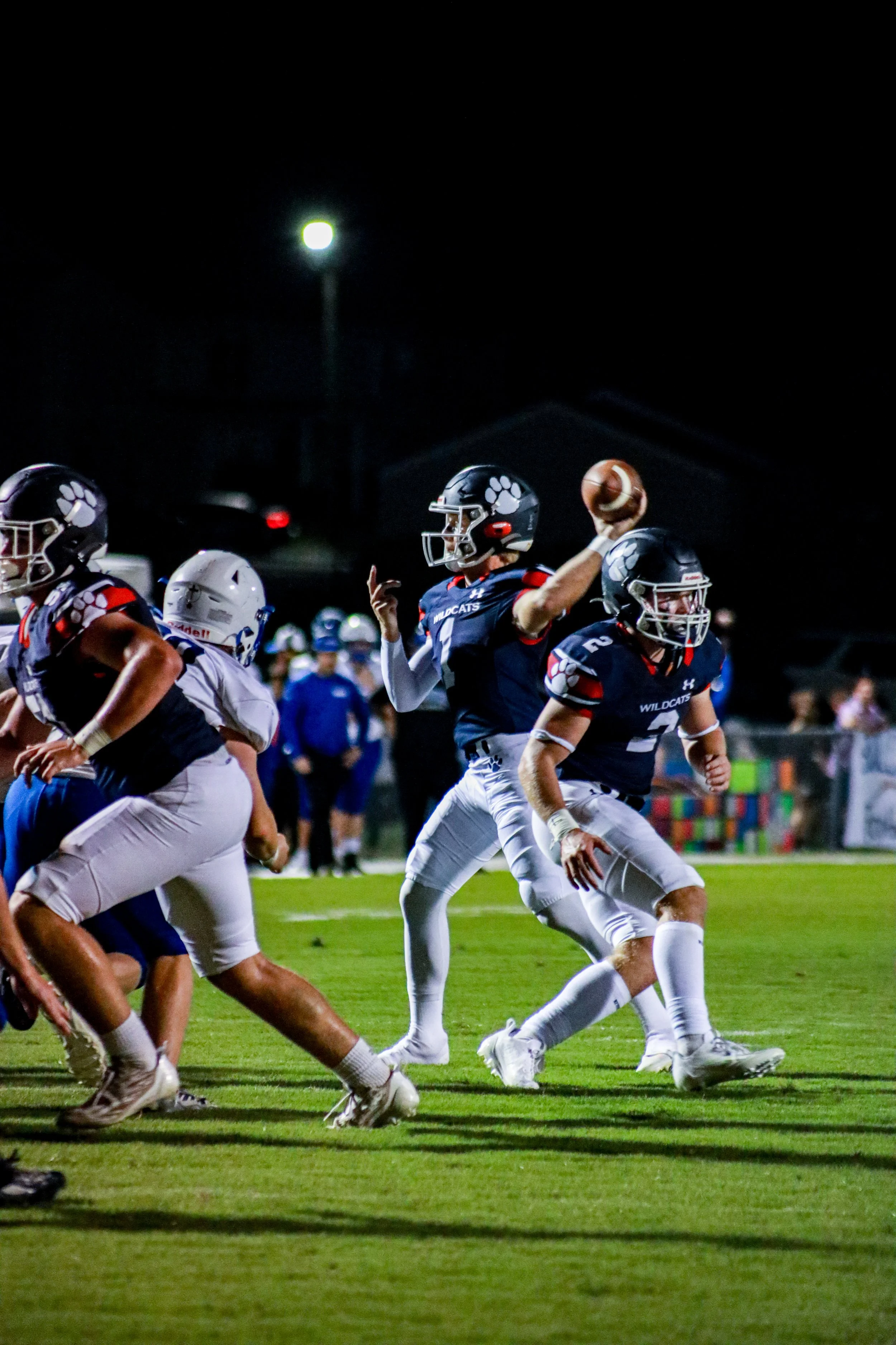 High school football quarterback in a Wildcats uniform throws a pass under stadium lights as teammates block defenders on the field.