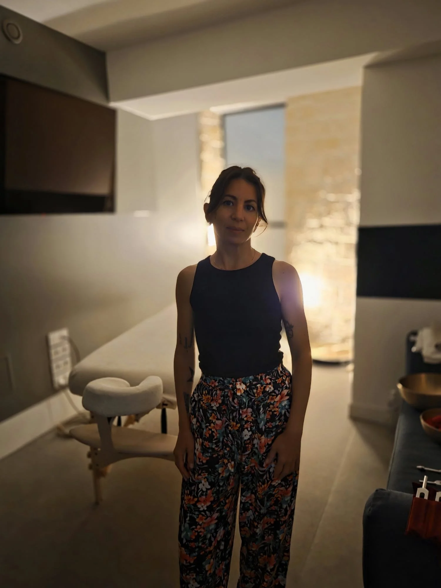 Une femme debout dans un intérieur avec un éclairage doux, portant un haut noir sans manches et un pantalon à fleurs.