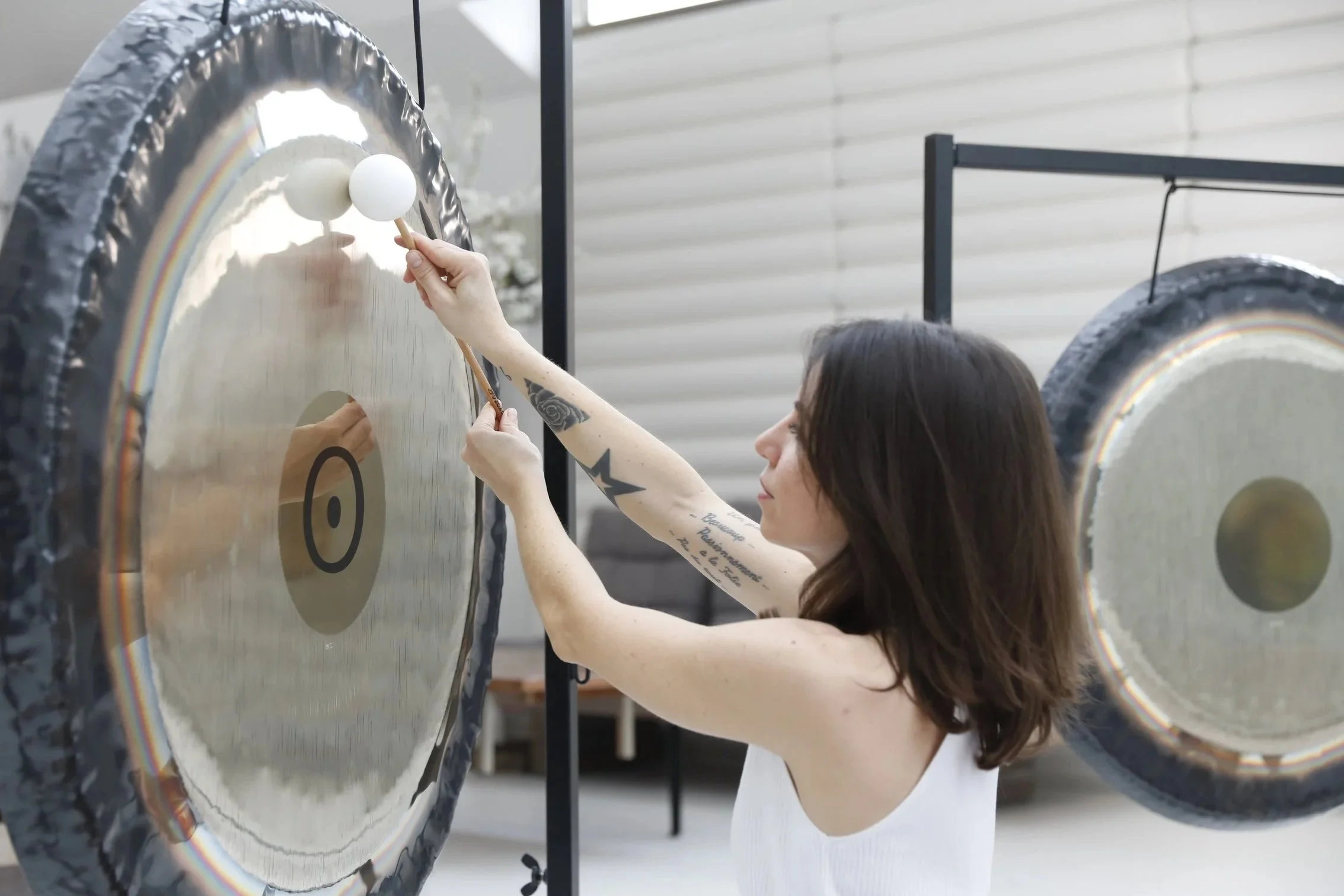 Une femme avec des tatouages joue du gong en utilisant deux maillets en bois avec des boules blanches, dans une salle lumineuse.