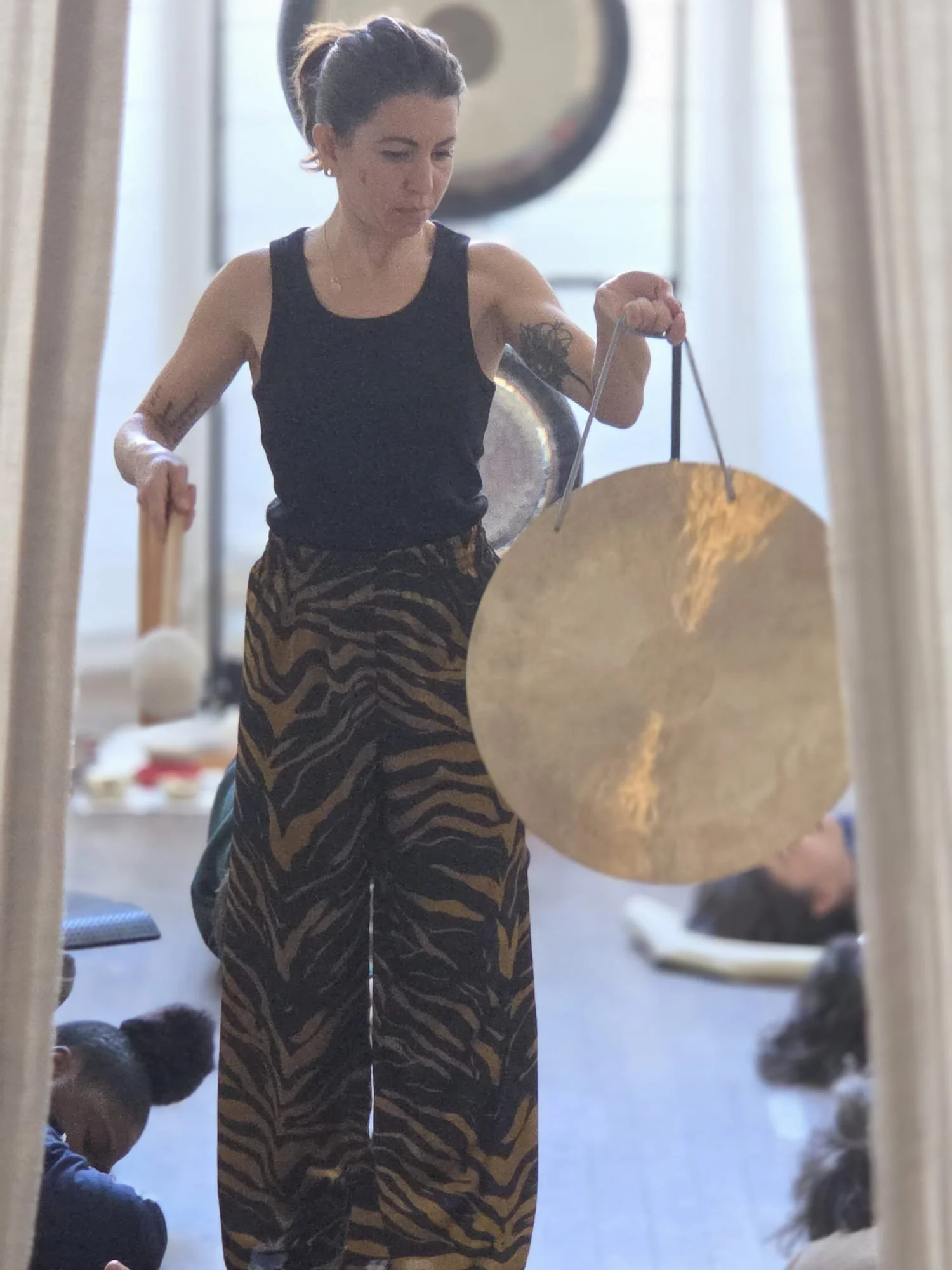 Une femme regardant dans un miroir, tenant un grand gong doré suspendu par une corde, portant un débardeur noir et un pantalon à motif zébré jaune et noir.