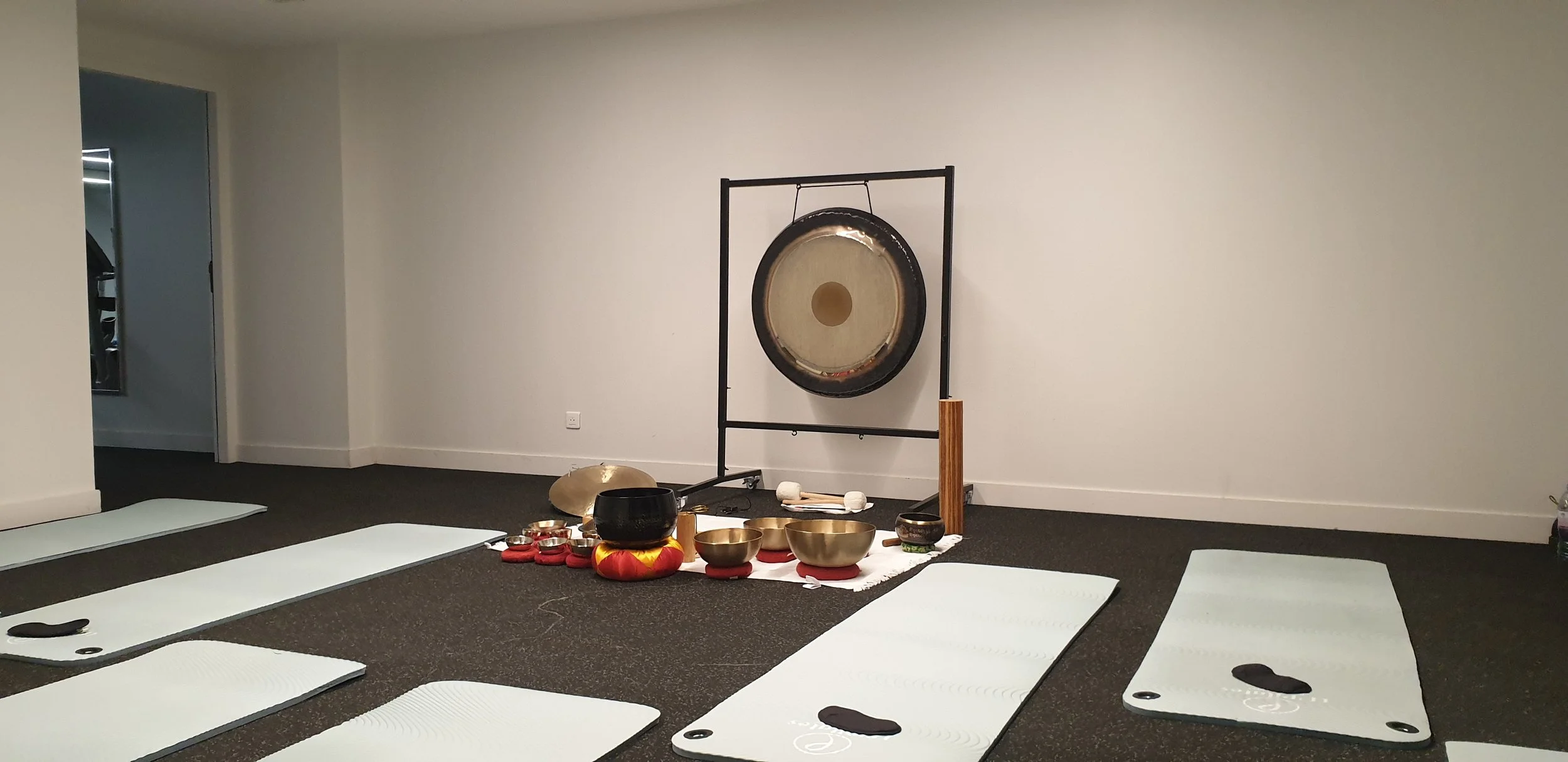 Salle de yoga ou méditation avec des tapis blancs, un gong suspendu sur un cadre noir, et divers bols tibétains disposés sur un tapis blanc au sol.