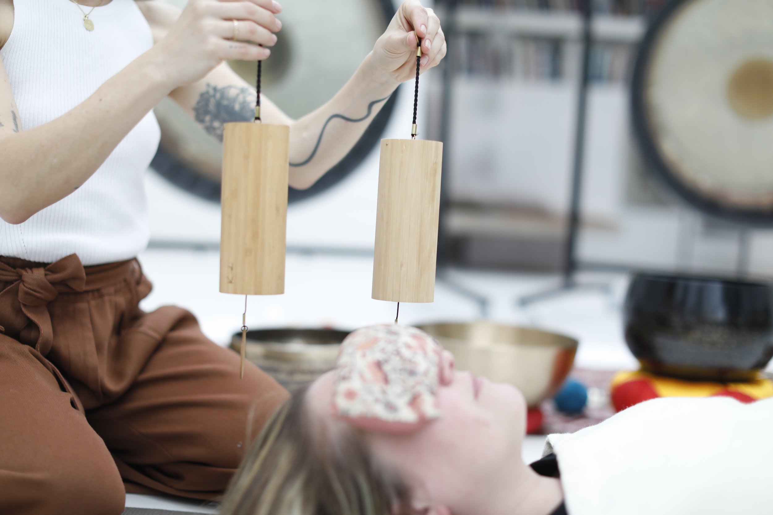 Une personne allongée, recevant un massage sonore avec deux bâtons en bois suspendus, avec des bols tibétains autour.