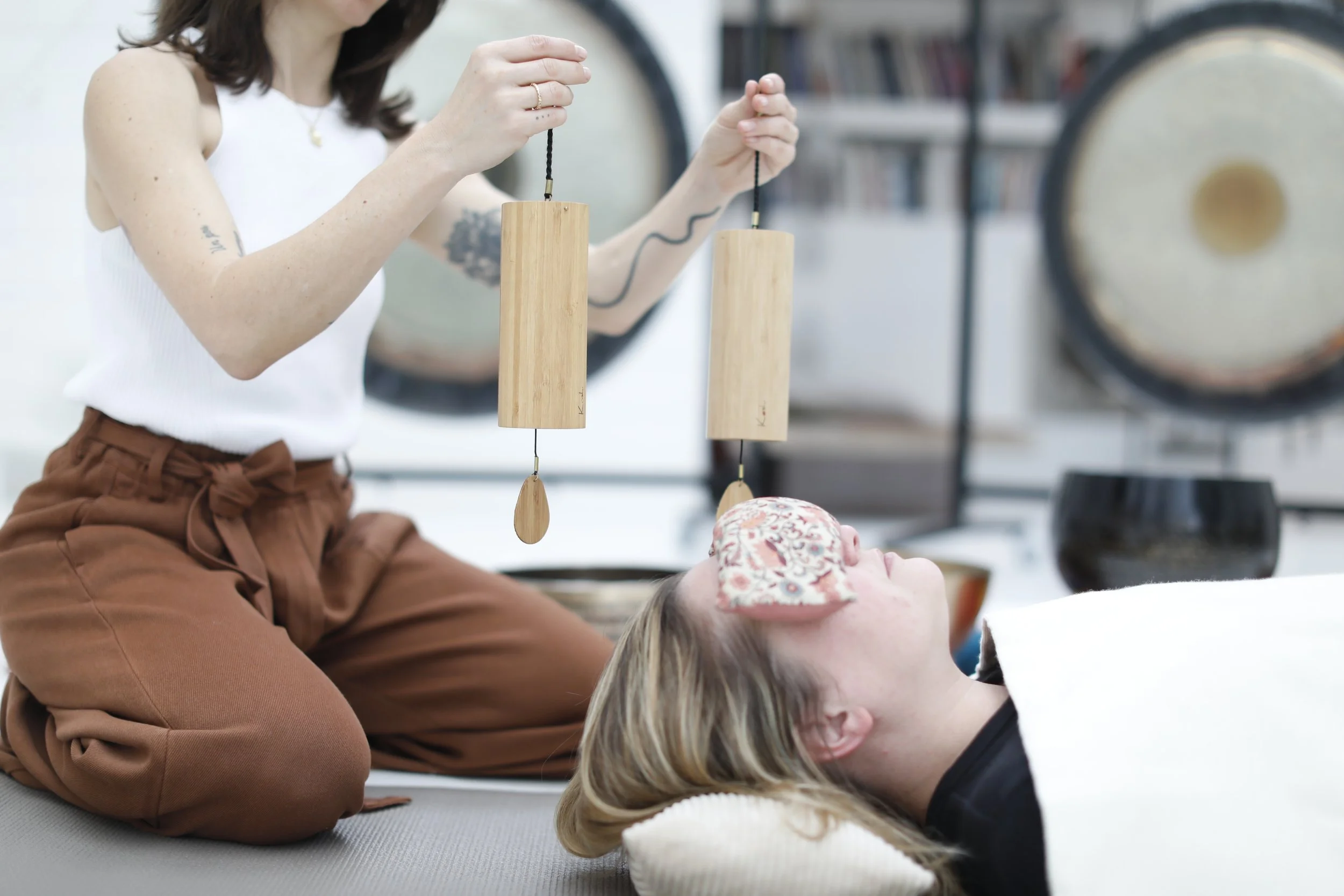 Une femme est allongée sur le sol avec un masque de coton sur le visage, tandis qu'une autre femme, assise à genoux, joue des instruments de percussion suspendus. L'arrière-plan montre des objets décoratifs et des livres.