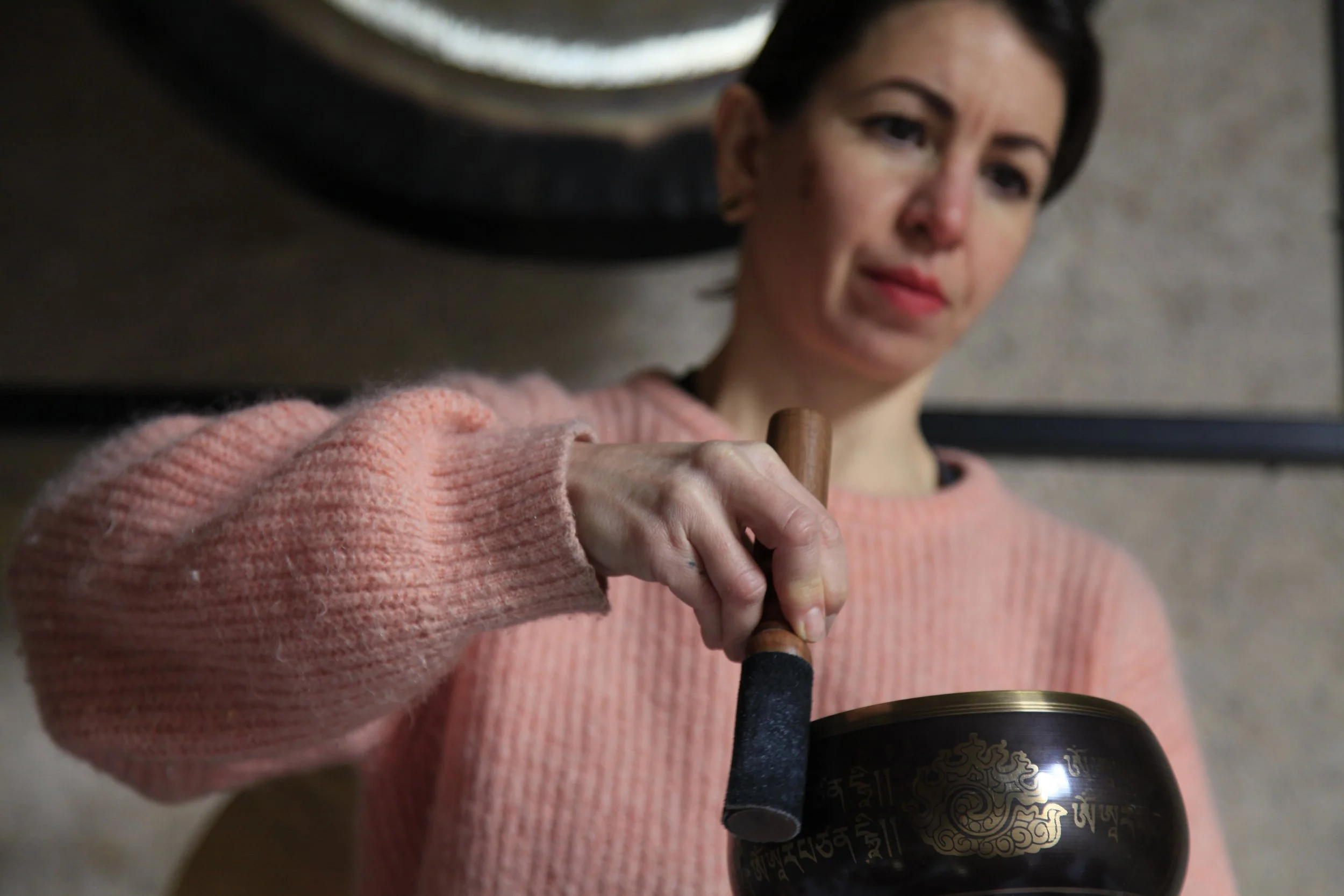 Une femme tient un bâton de percussion en bois et frappe un bol chantant tibétain en métal noir avec des inscriptions dorées, dans un intérieur.