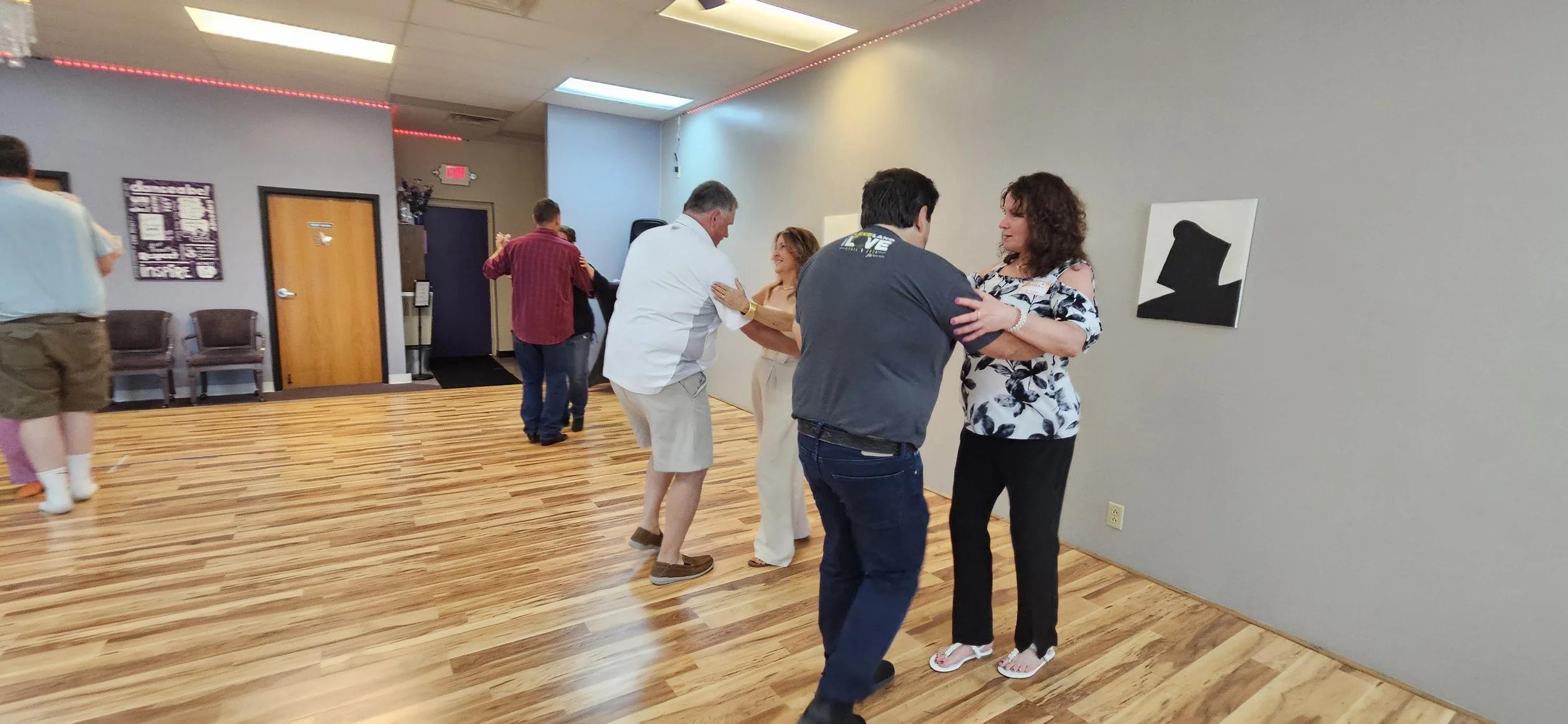 People dancing in pairs inside a room with wooden floors and gray walls.