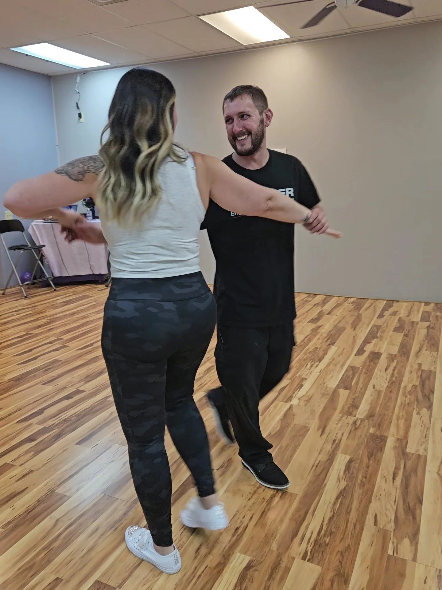 A man and woman are dancing together in a room with wooden flooring and plain walls, smiling and looking at each other.
