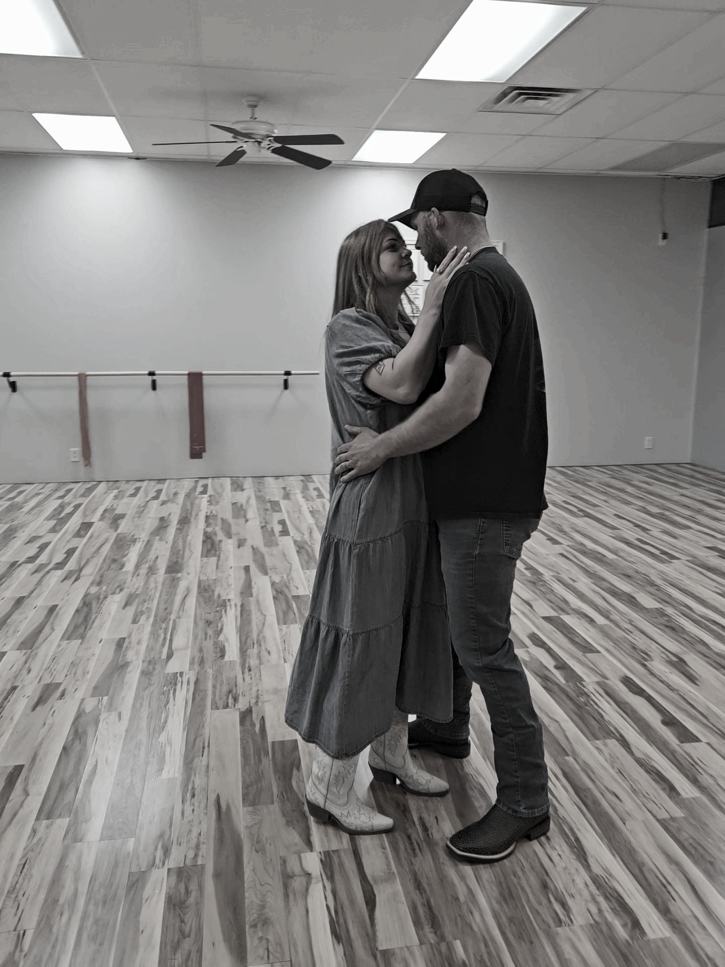 A couple dancing closely in a dance studio with wooden flooring, plain walls, and a ballet barre in the background.