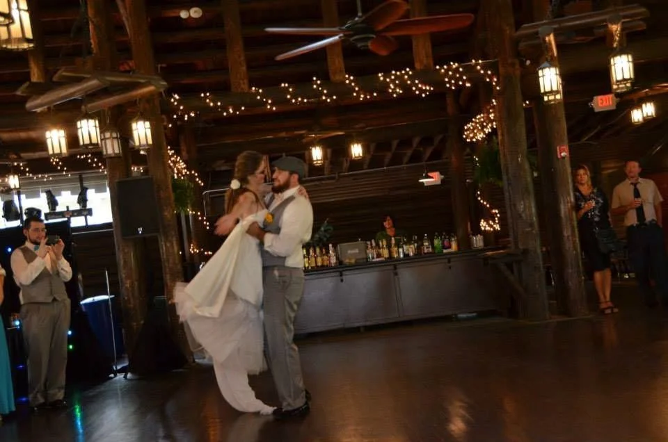 A bride and groom dance together at their wedding reception in a rustic wooden venue decorated with string lights. A DJ booth and bar are visible in the background, with guests watching and taking photos.