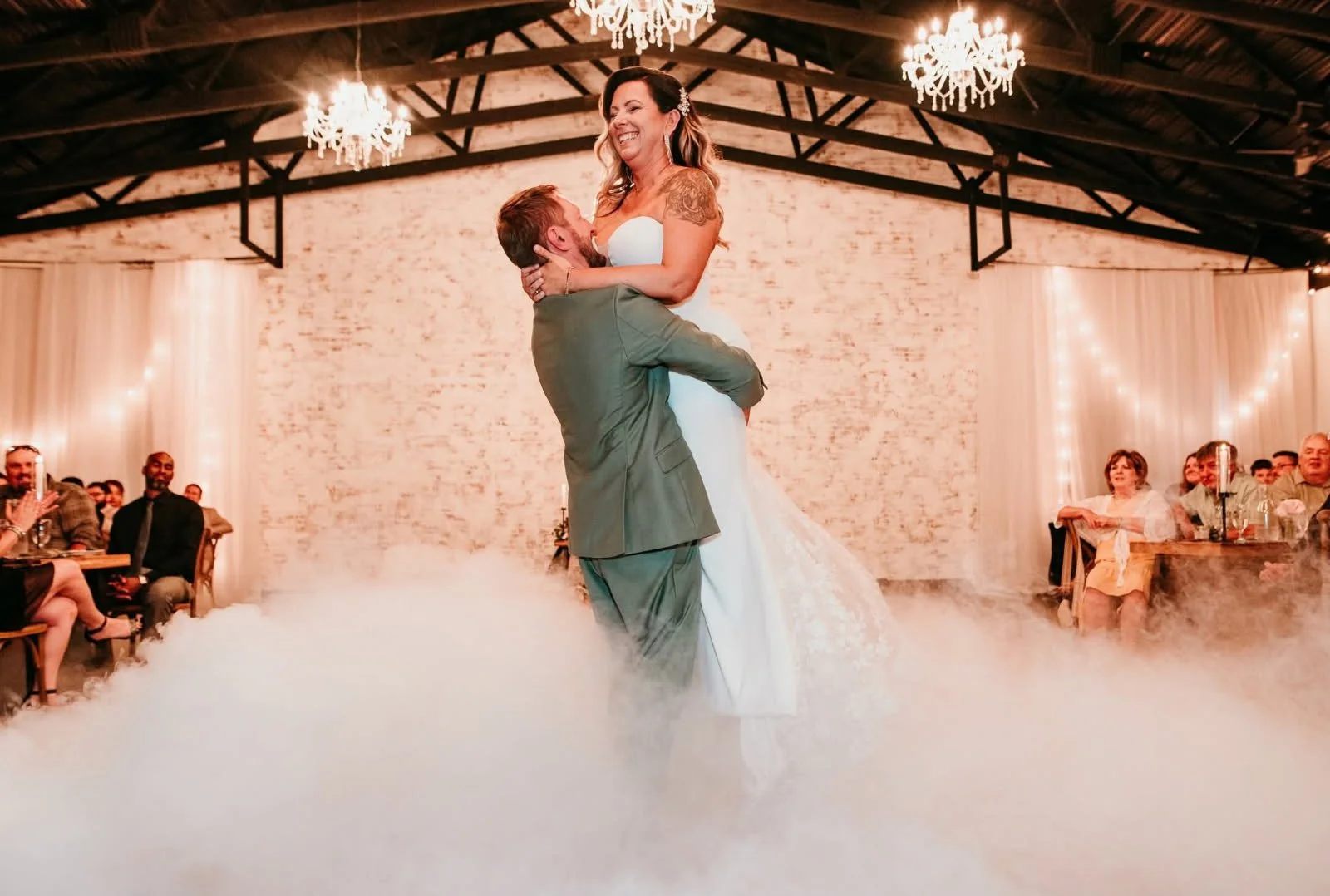 A bride and groom dancing during their wedding reception, with guests watching, in a rustic venue decorated with chandeliers, white drapery, and string lights.