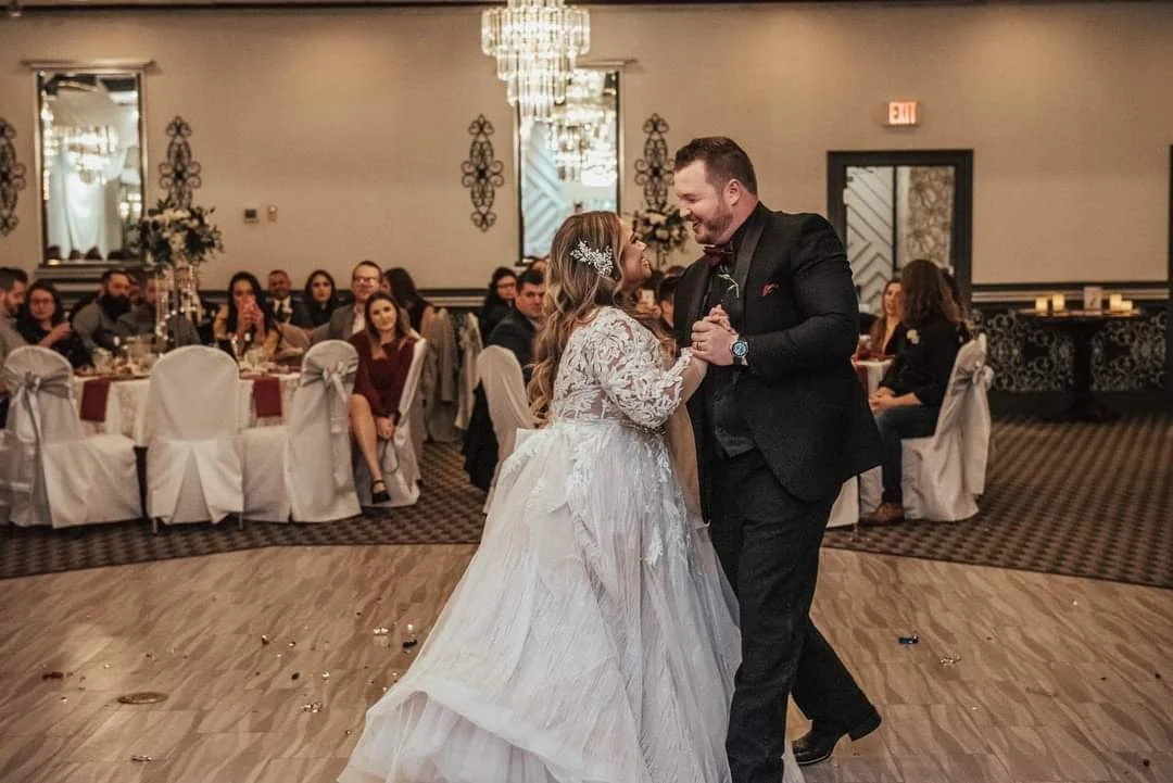 A bride and groom dance together at their wedding reception, surrounded by seated guests in an elegantly decorated banquet hall.