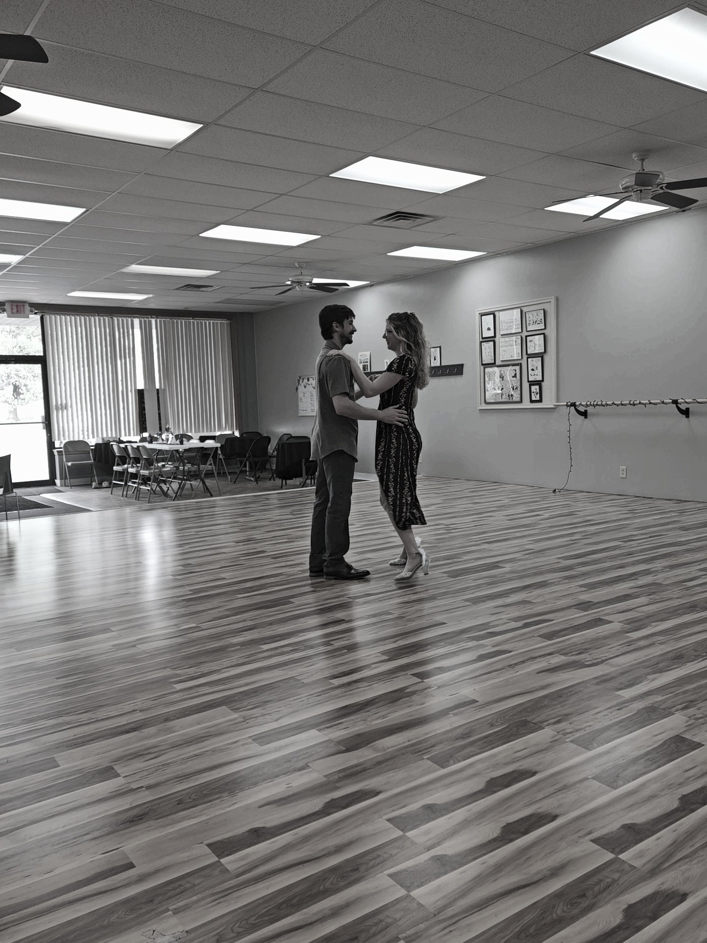 A couple dancing in an empty dance studio with wood floors, framed artwork on the wall, and a large window with vertical blinds.