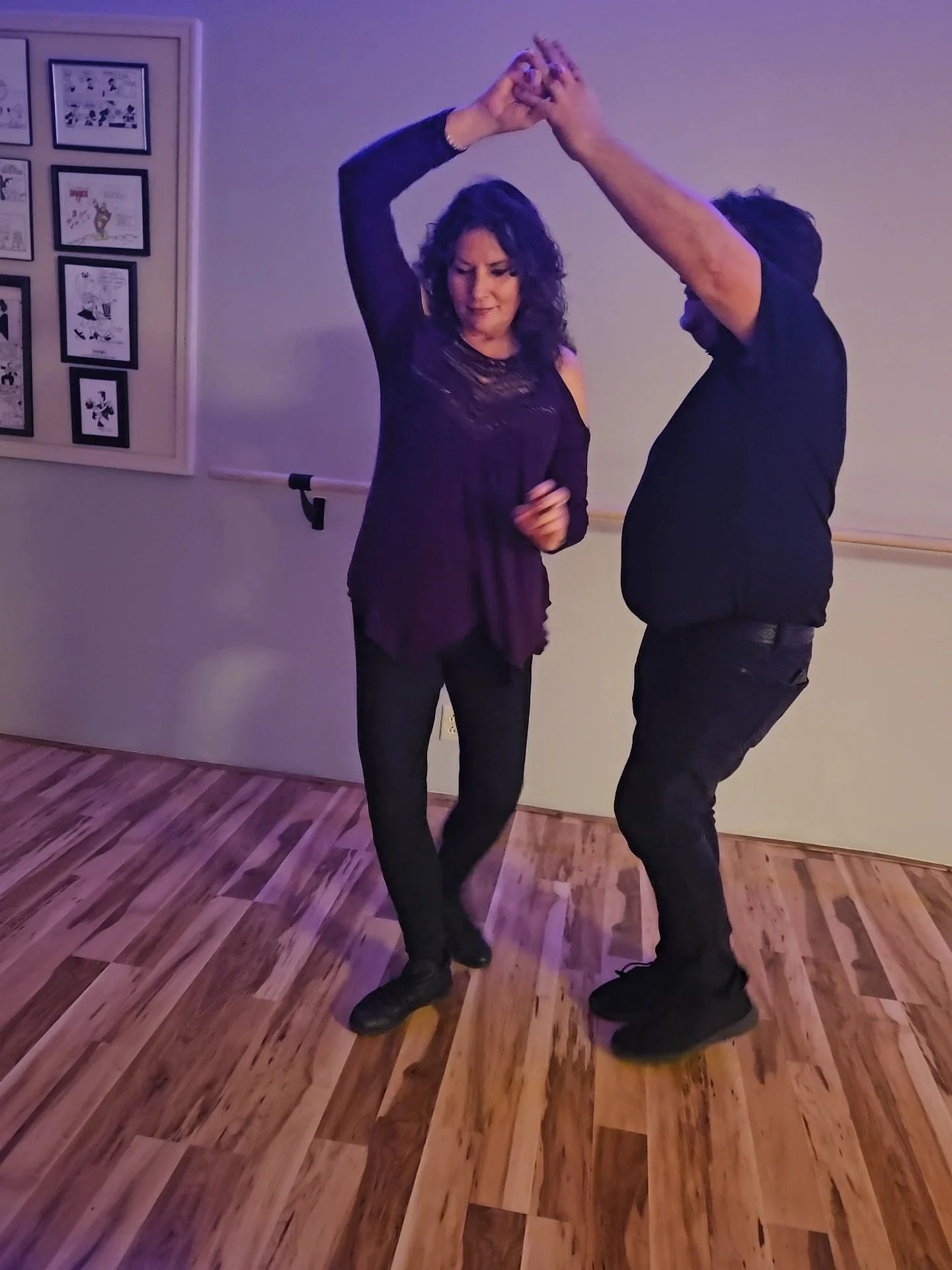 A man and woman dancing together indoors on a wooden floor, with framed pictures on the wall behind them.