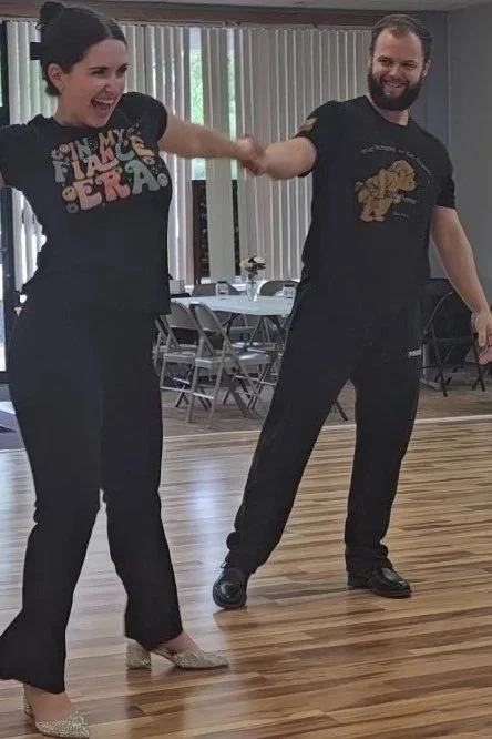 A woman and man dancing together indoors, holding hands, with a wooden floor and tables with chairs in the background.
