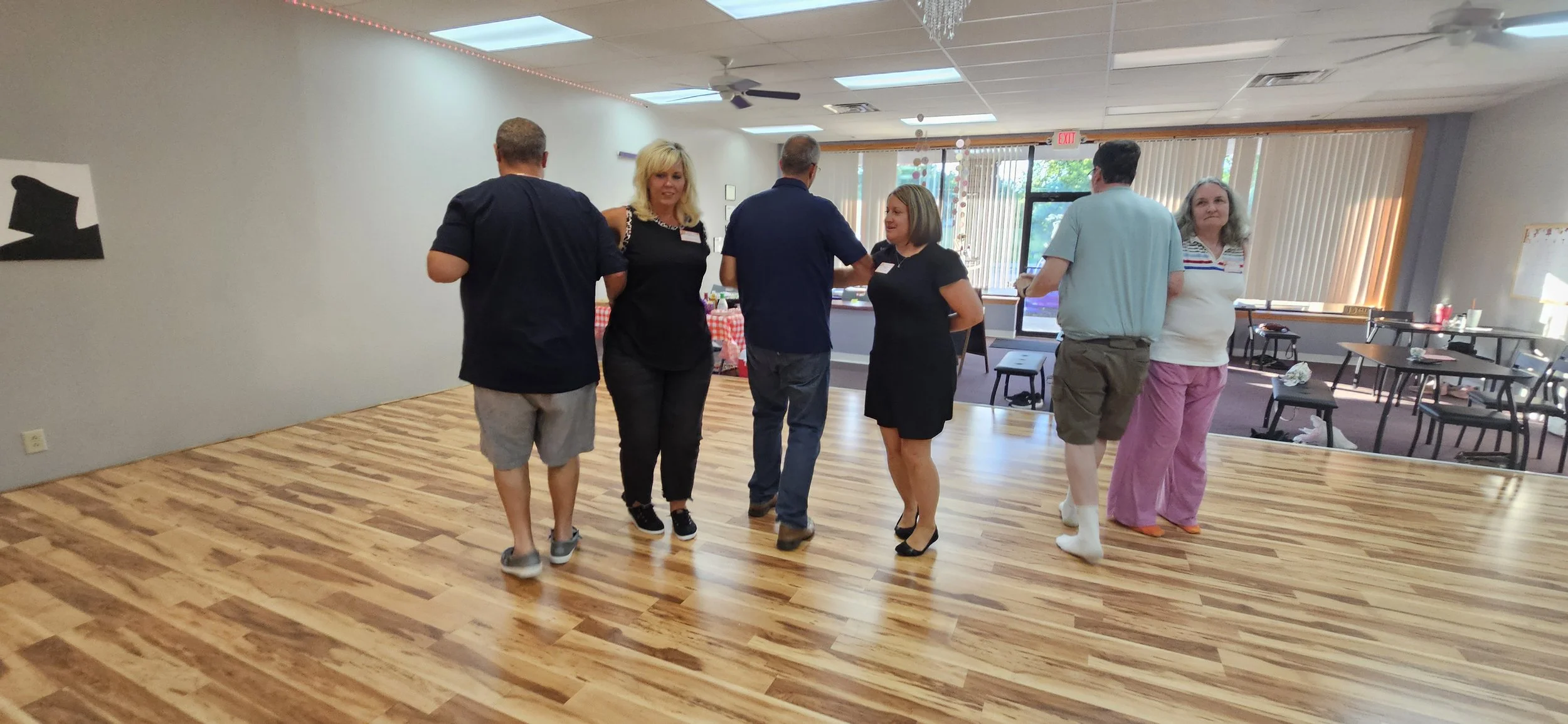 Six people dancing in a room with wooden flooring, casual and semi-formal attire, and large windows letting in natural light.