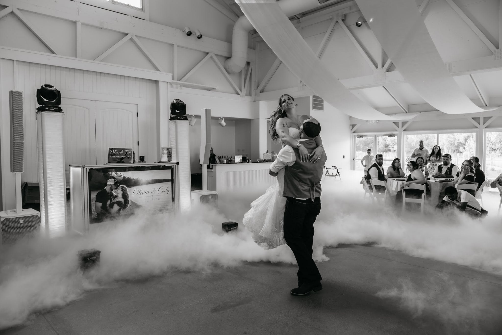 A bride and groom are dancing at their wedding reception in a bright, spacious hall with large windows, guests sitting at tables, and a display with the couple's names and wedding date.