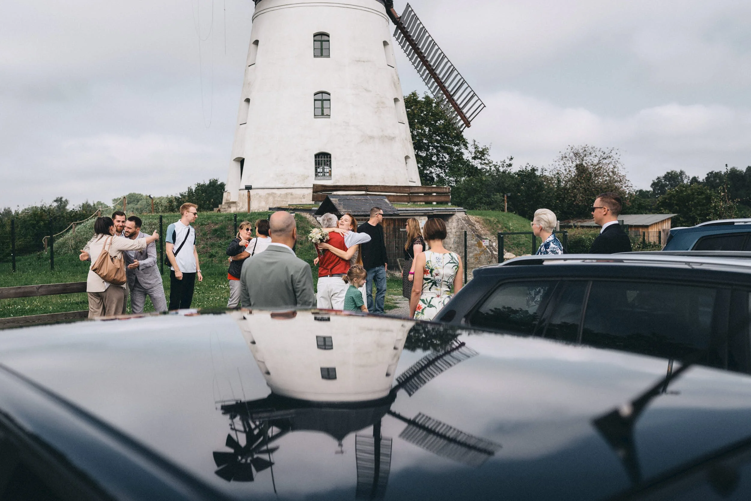 Hochzeitsparty im Freien vor einer weißen Windmühle, einige Gäste umarmen sich, andere unterhalten sich, und ein Baby ist sichtbar, während im Vordergrund die reflektierende Oberfläche eines Autos die Szene spiegelt.