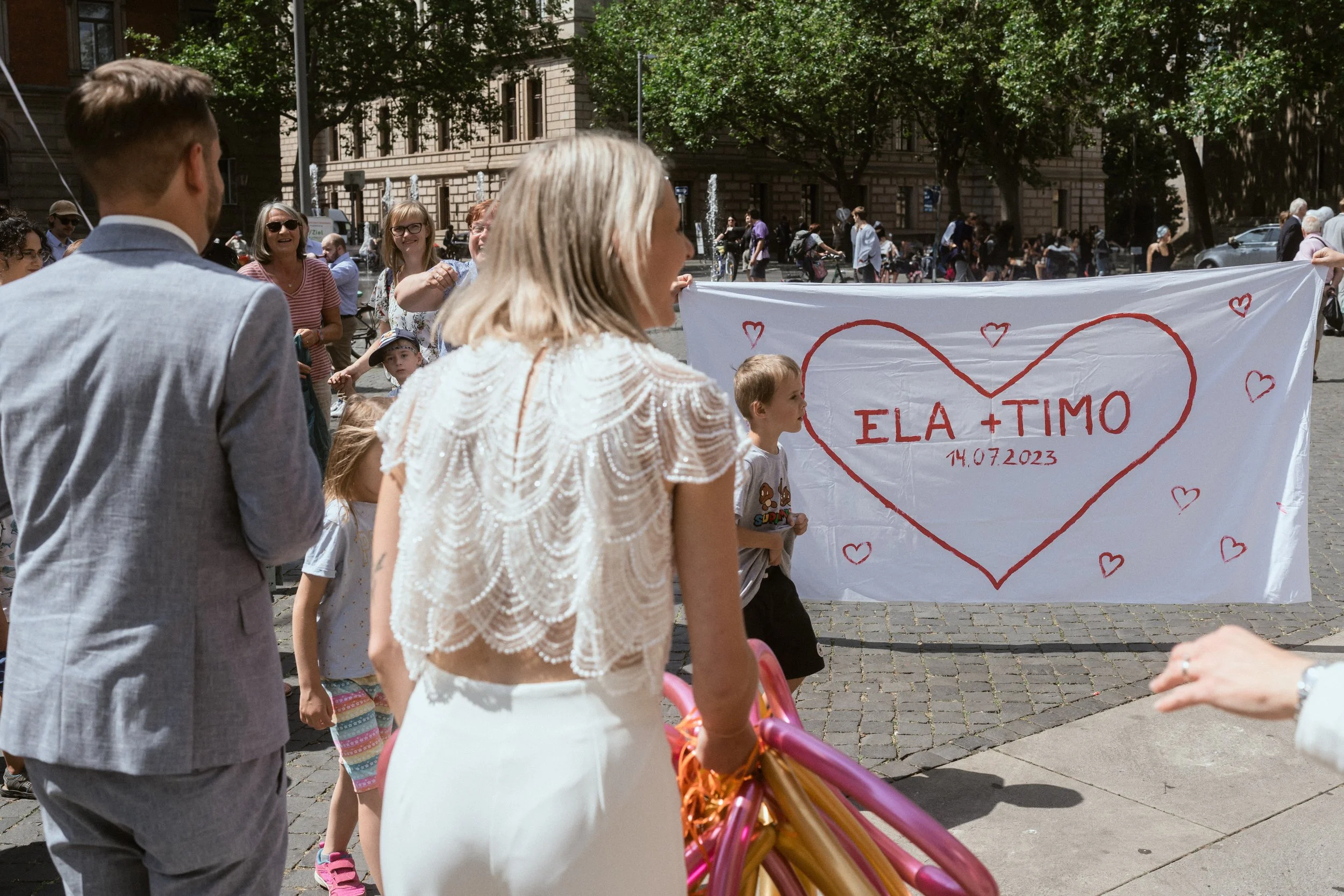 Menschen bei einer Hochzeitsfeier im Freien, eine Person hält ein Banner mit Herz und den Namen ELA + Timo, Datum 14.07.2023.