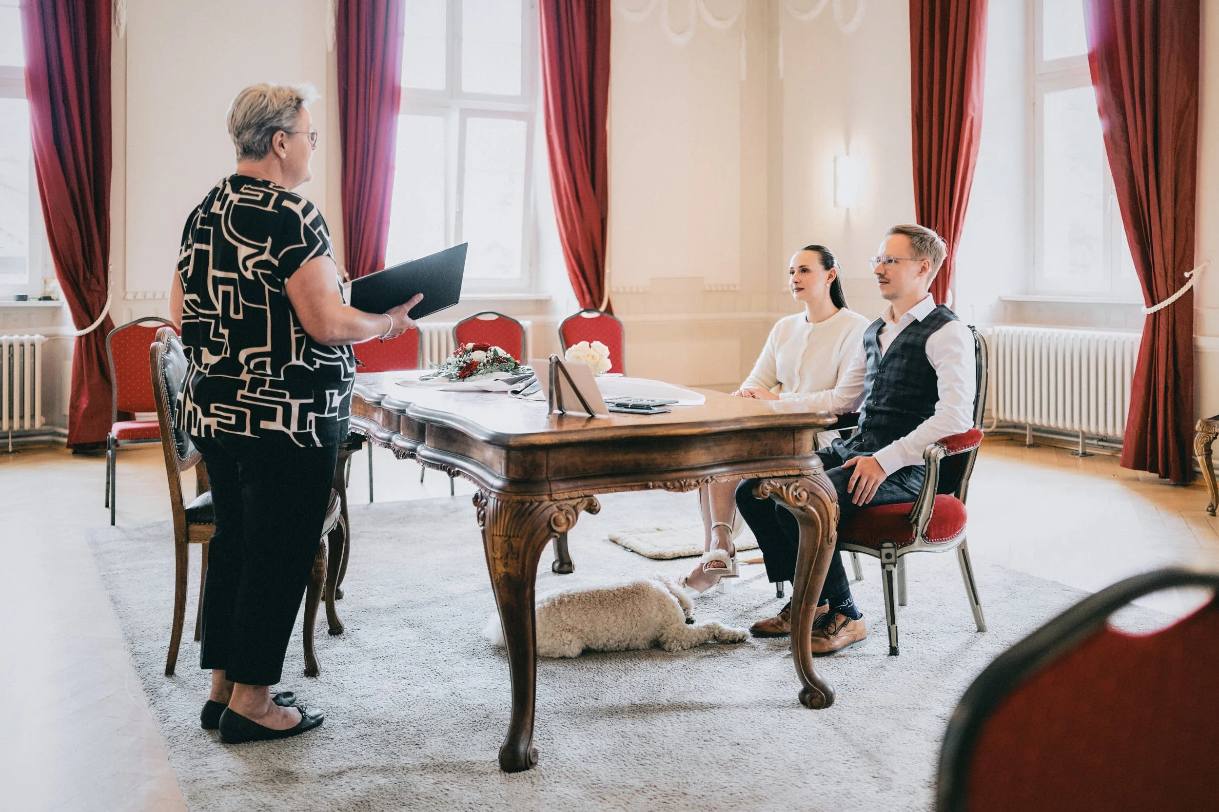 Eine Frau steht vor einem Tisch, während zwei Personen auf einem Stuhl sitzen, in einem eleganten Raum mit hohen Fenstern und roten Vorhängen.