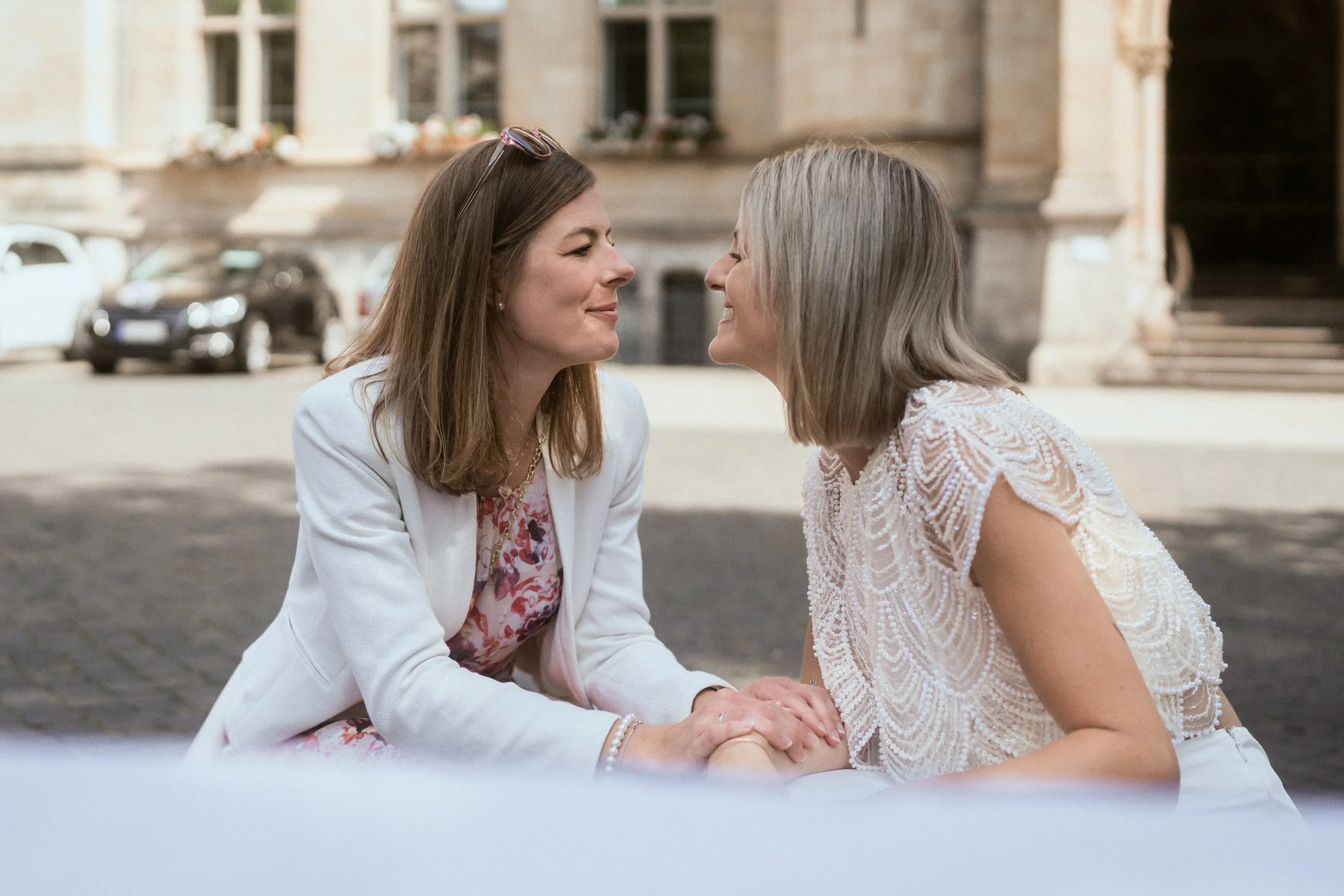Zwei Frauen sitzen sich bei einem Händedruck gegenüber. Sie lachen und schauen sich liebevoll an. Im Hintergrund eine alte Gebäude Fassaden mit Blumen in Fensterkästen.