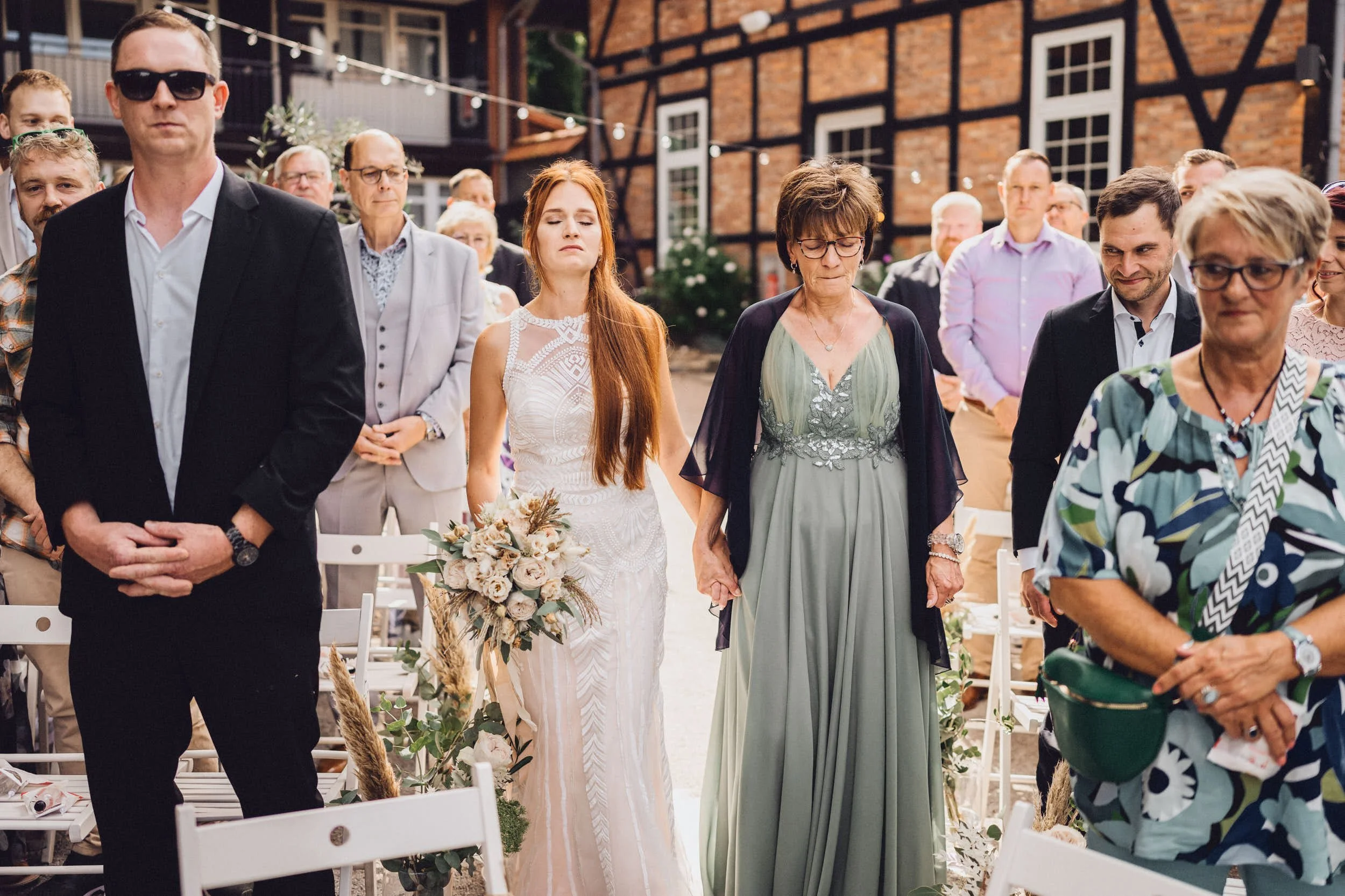 Wedding ceremony with guests standing, a bride in a white dress holding a bouquet, and a woman in a green dress, outdoors with a brick building background.