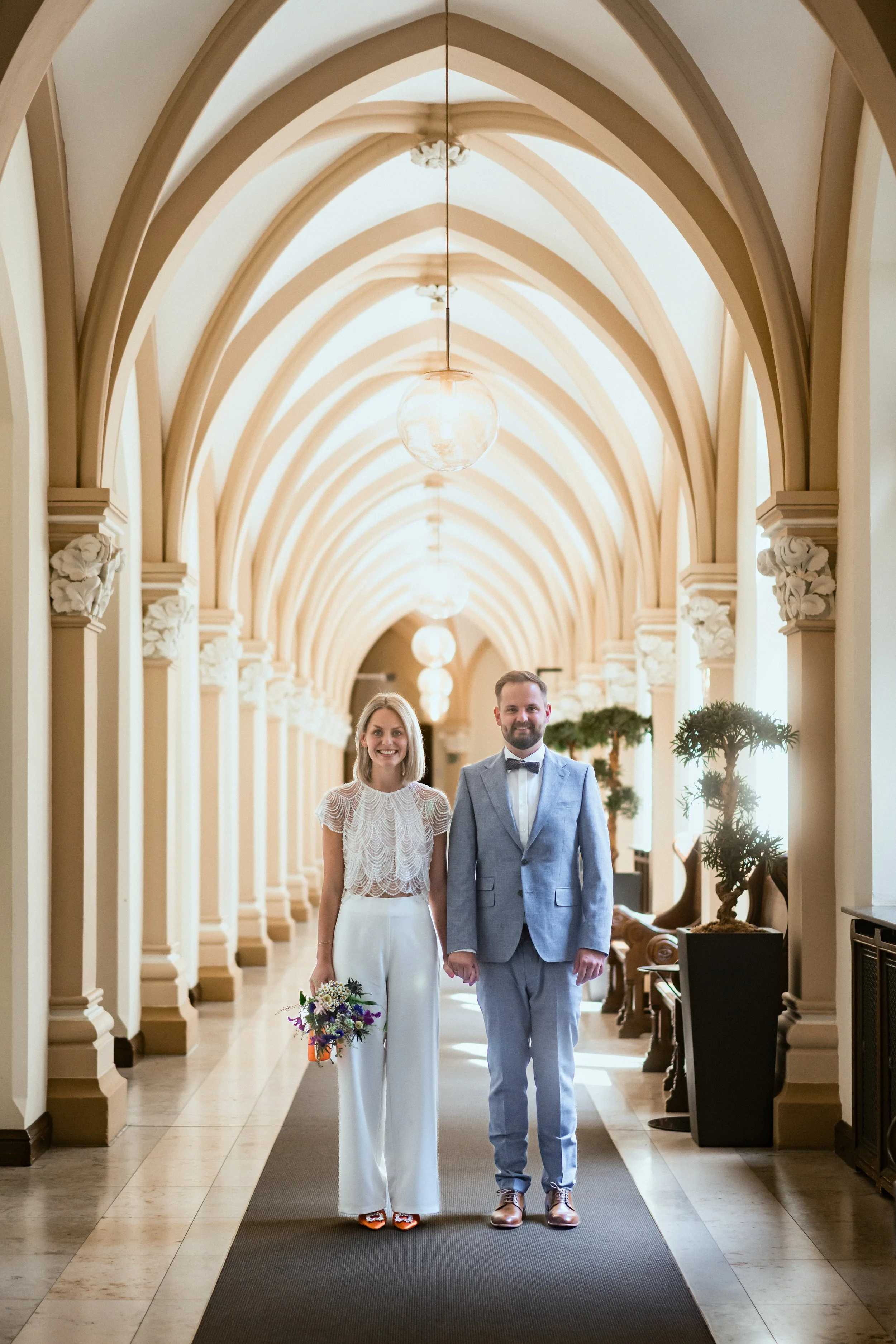 Ein glückliches Paar, ein Mann in einem grauen Anzug und eine Frau in einem weißen Hochzeitsoutfit, schlendern durch einen eleganten Flur mit hohen Gewölbedecken, Pflanzen und dekorativen Lampen.