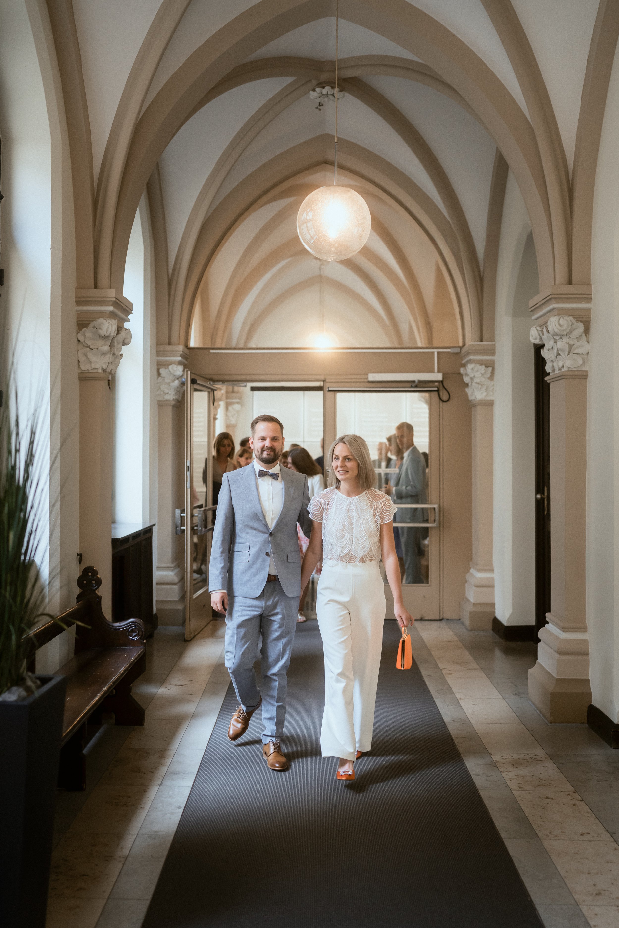 Ein Mann und eine Frau gehen Hand in Hand durch einen Flur in einem eleganten Gebäude, vermutlich nach einer Hochzeit.
