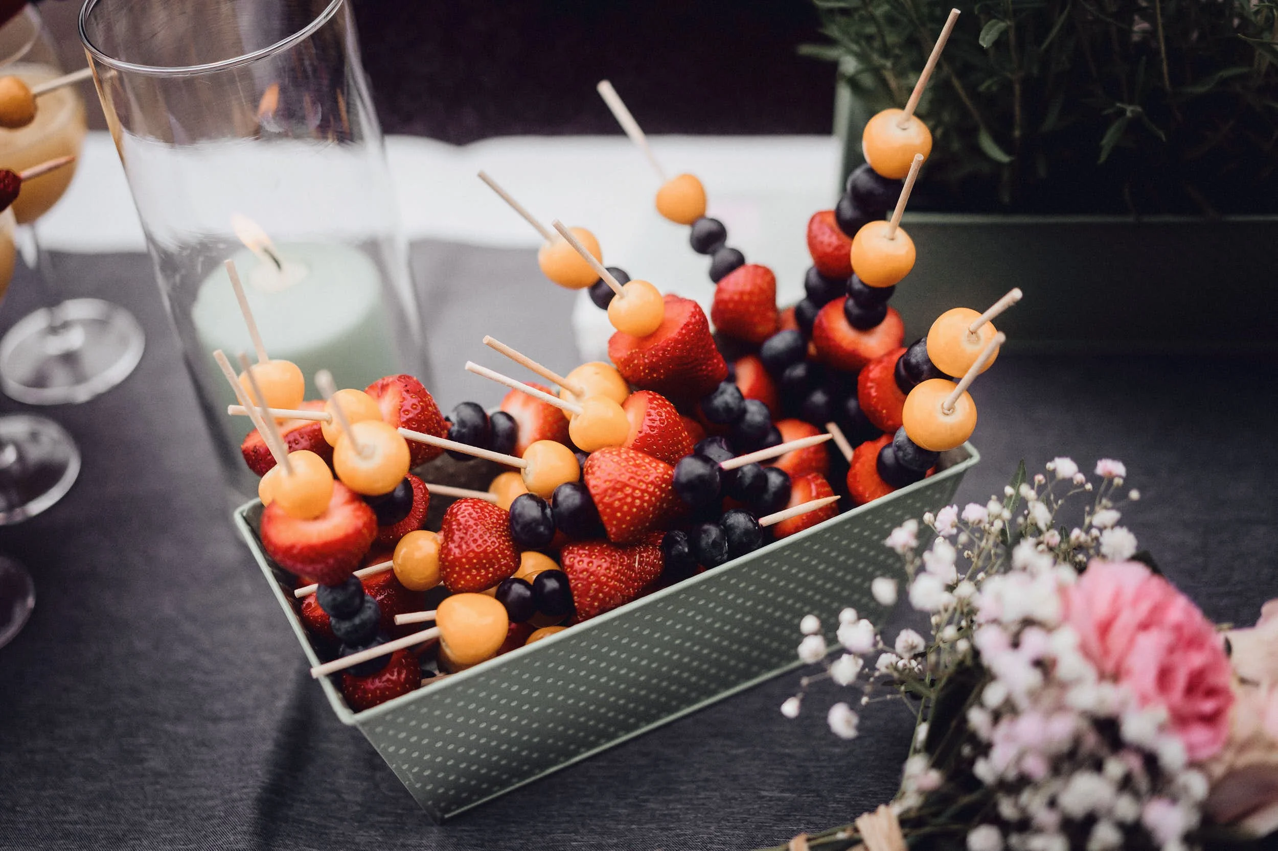 Obstspieße mit Erdbeeren, Blaubeeren und Honigmelone in einem Behälter, umgeben von einem brennenden Kerzenständer und Blumen.