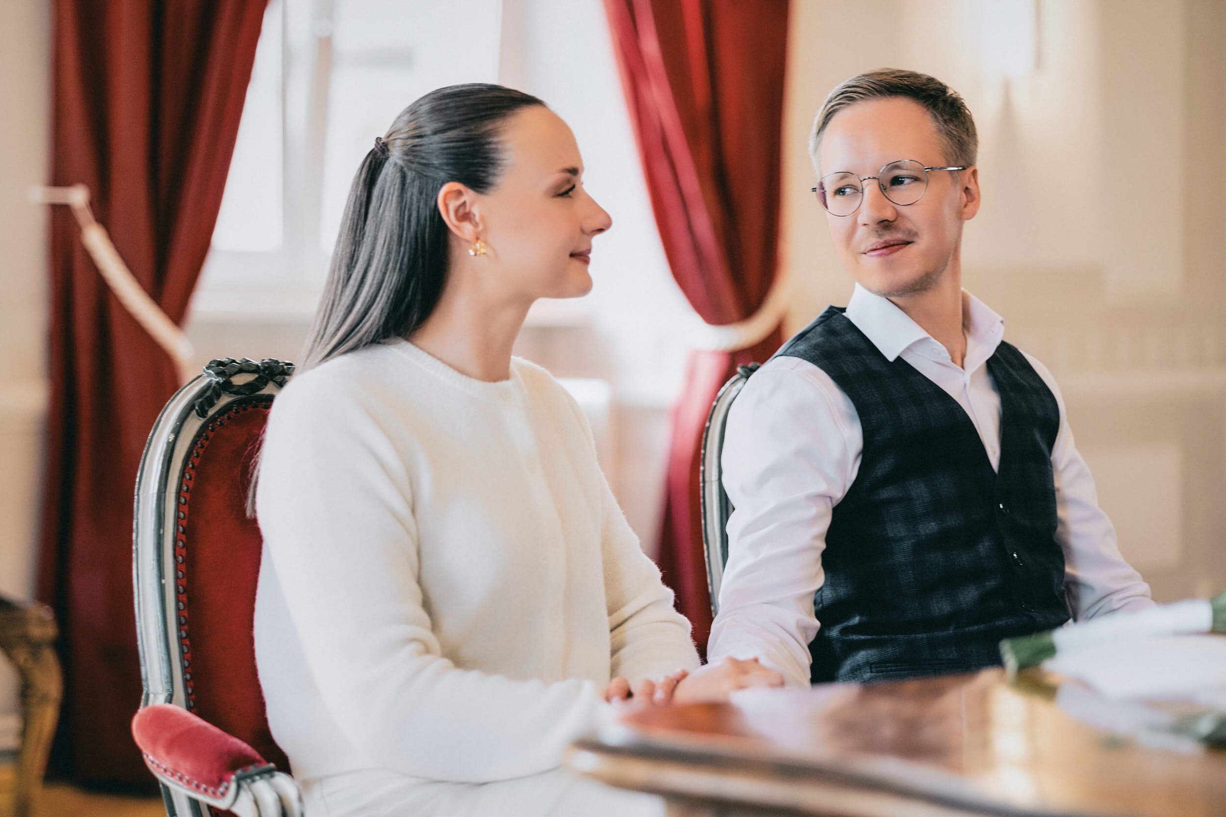 Ein Mann und eine Frau sitzen nebeneinander an einem Tisch in einem eleganten Raum mit roten Vorhängen. Sie scheinen sich zu unterhalten.