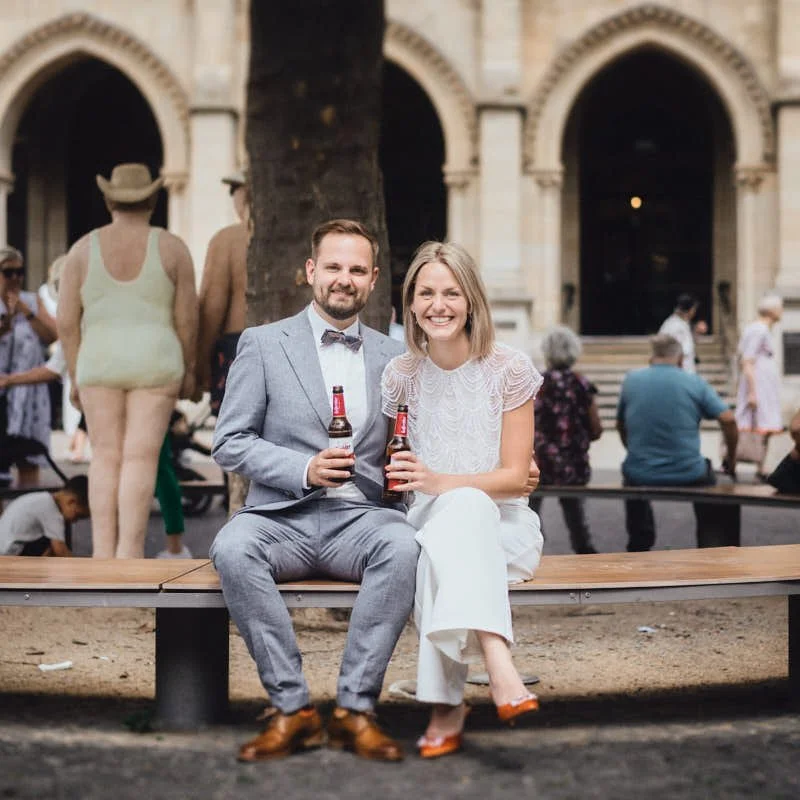 Ein Mann in einem grauen Anzug und eine Frau in weißem Kleid sitzen auf einer Bank, lachen und halten jeweils eine Flasche Bier. Im Hintergrund sind Menschen, ein Baum und eine historische Gebäude-Architektur.