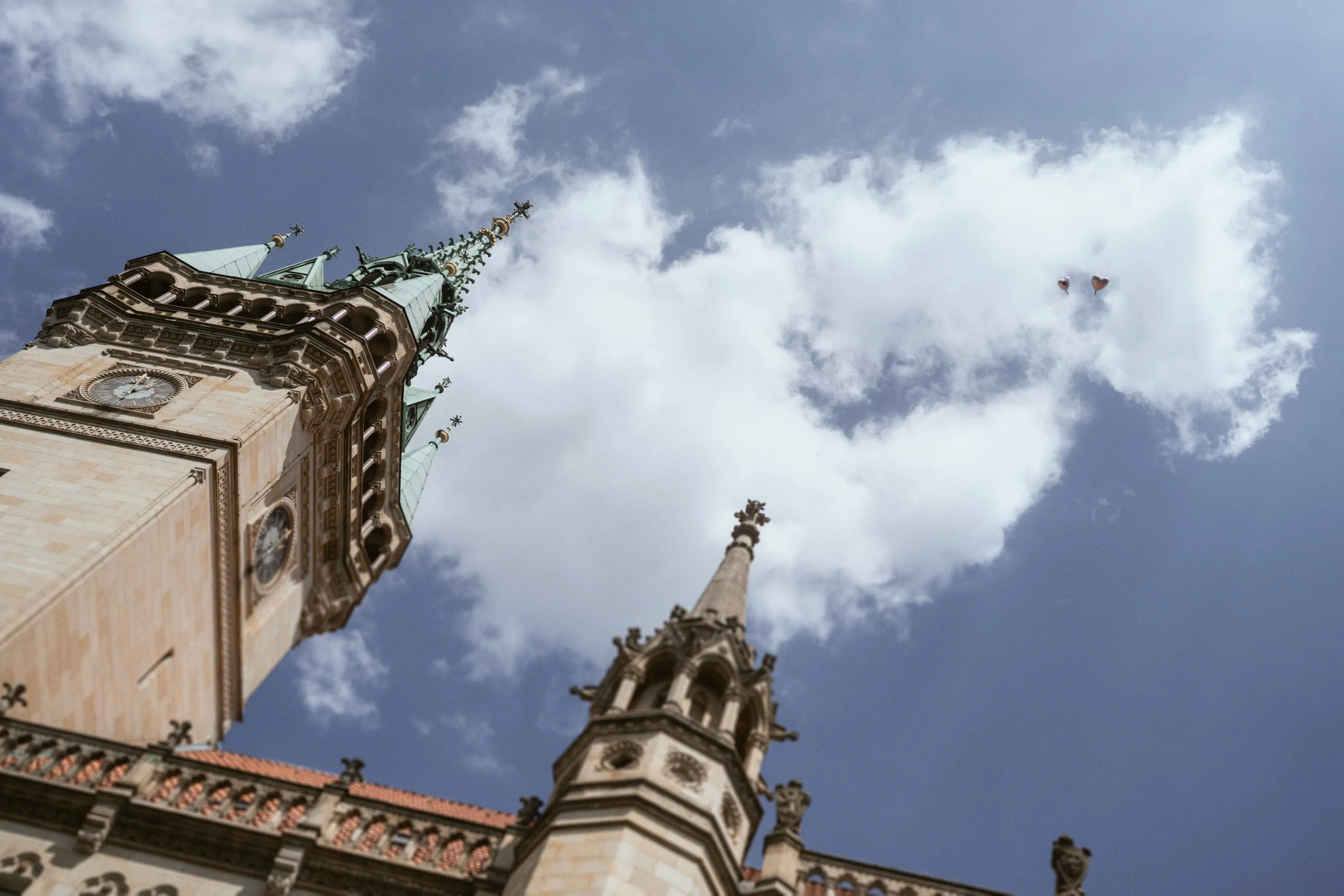 Blick nach oben auf den Kirchturm einer gotischen Kirche mit Himmel und Wolken im Hintergrund, inklusive Kugeln und Herzballons