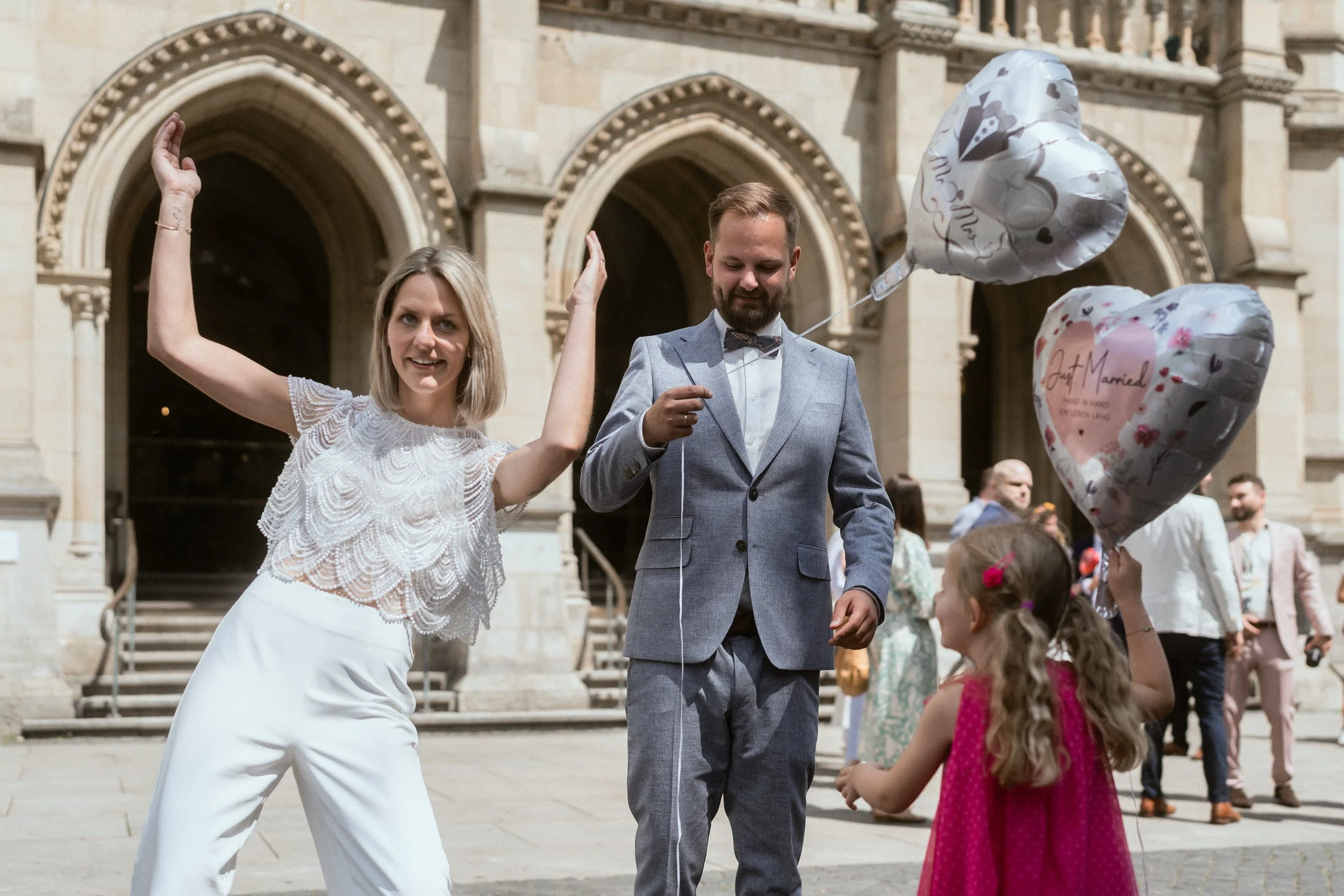 Ein Hochzeitsfest vor einem historischen Gebäude. Ein Brautpaar steht in der Mitte, umgeben von Kindern und Gästen. Das Brautpaar hält Ballons, eine hat den Text "Just Married".