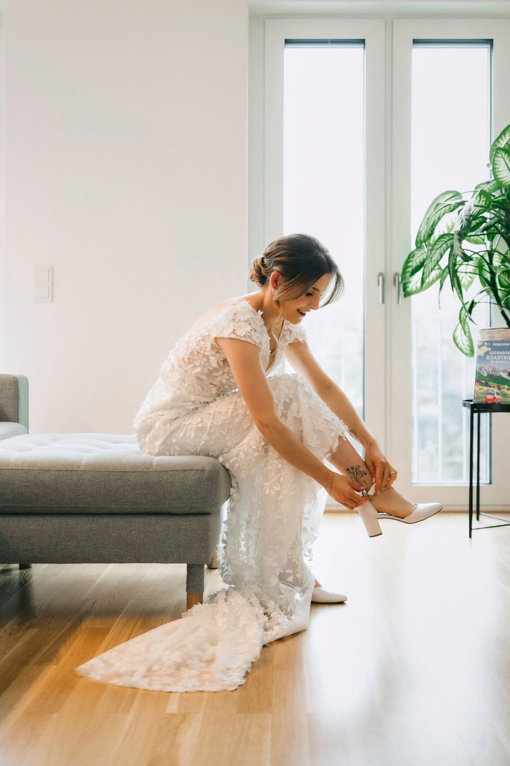 Eine Frau im weißen Hochzeitskleid sitzt auf einer Bank und steckt sich weiße High Heels an, im Hintergrund große Fenster und ein grüner Pflanze.