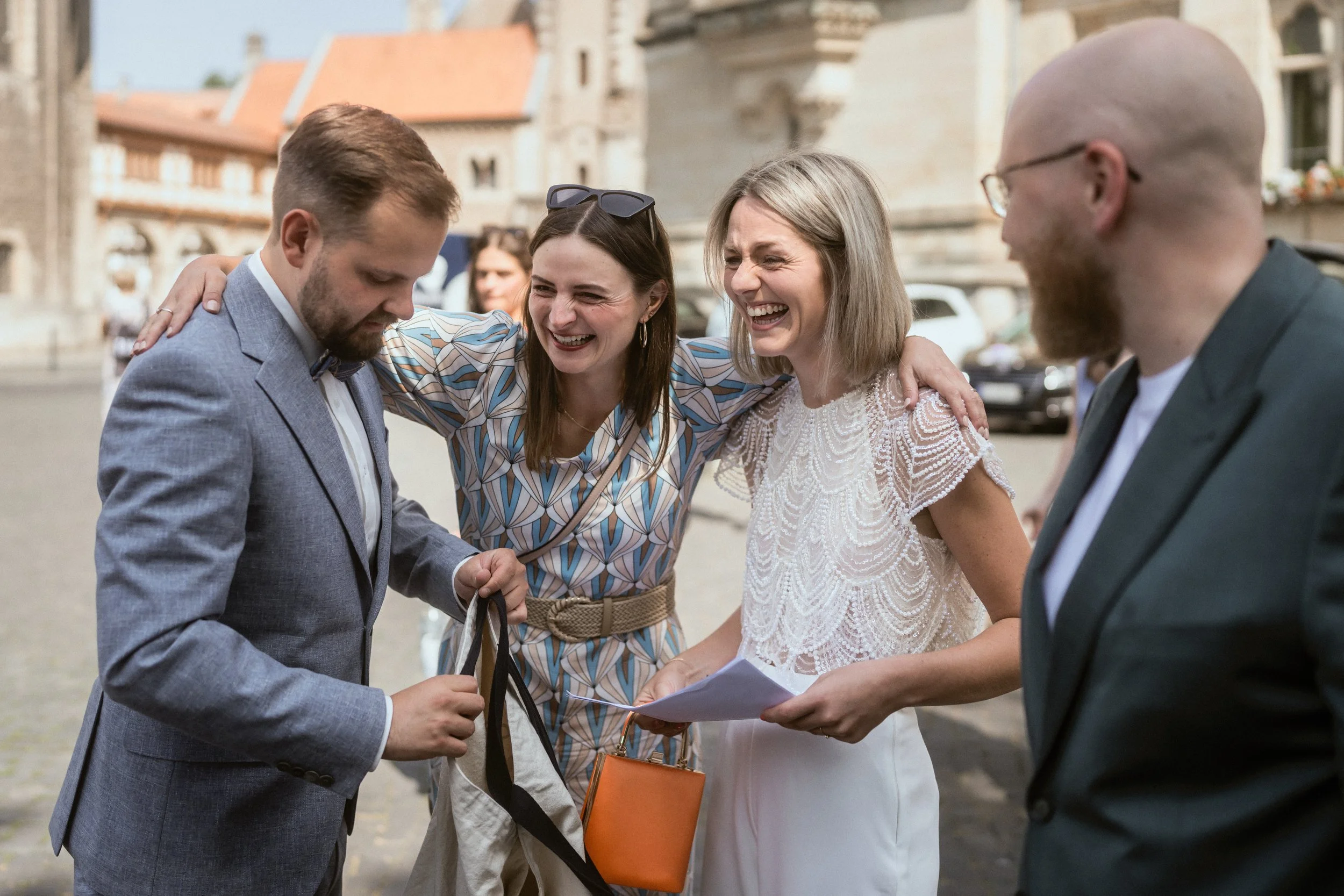Vier Menschen, zwei Männer und zwei Frauen, lachen und sprechen miteinander im Freien, während sie Unterlagen und eine Tasche halten.
