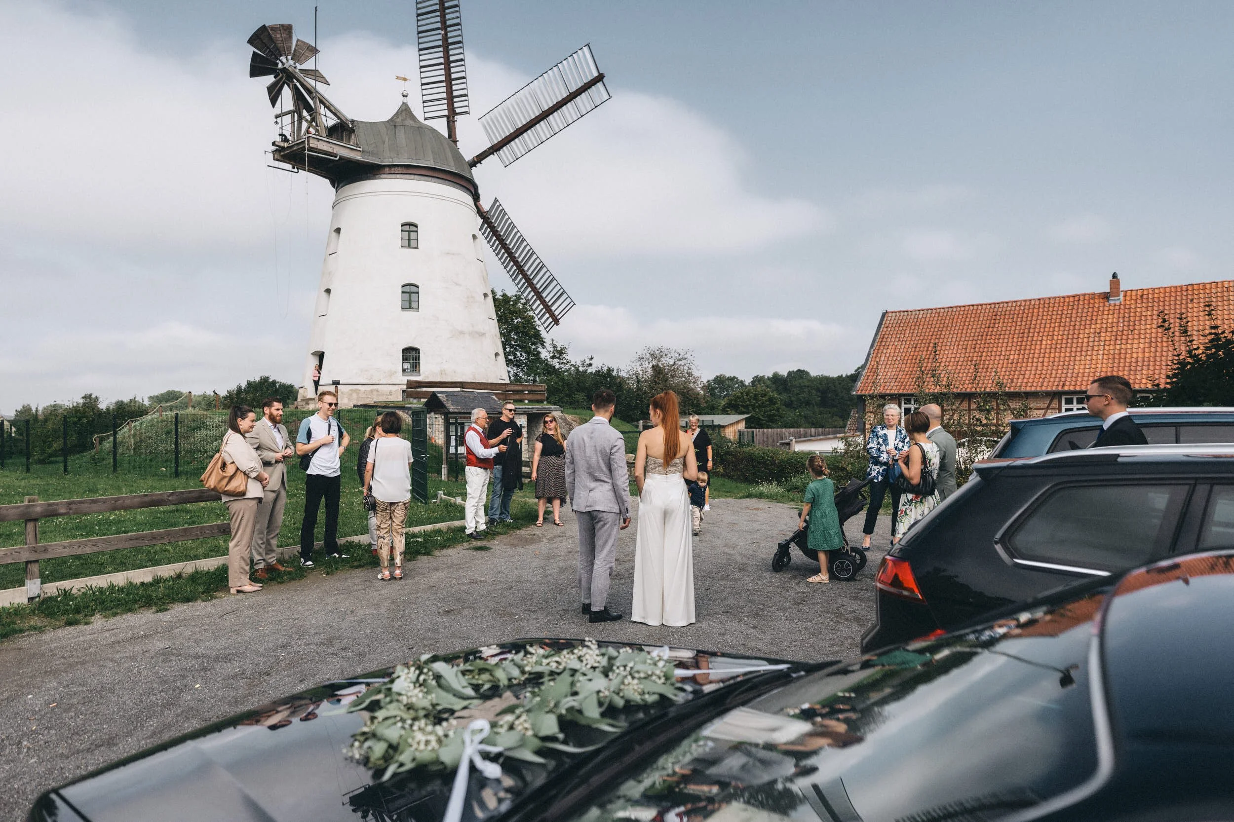Eine Gruppe von Menschen versammelt sich im Freien vor einem weißen Windmühlenturm, möglicherweise bei einer Hochzeit oder Veranstaltung, während das Auto im Vordergrund mit Blumen geschmückt ist.