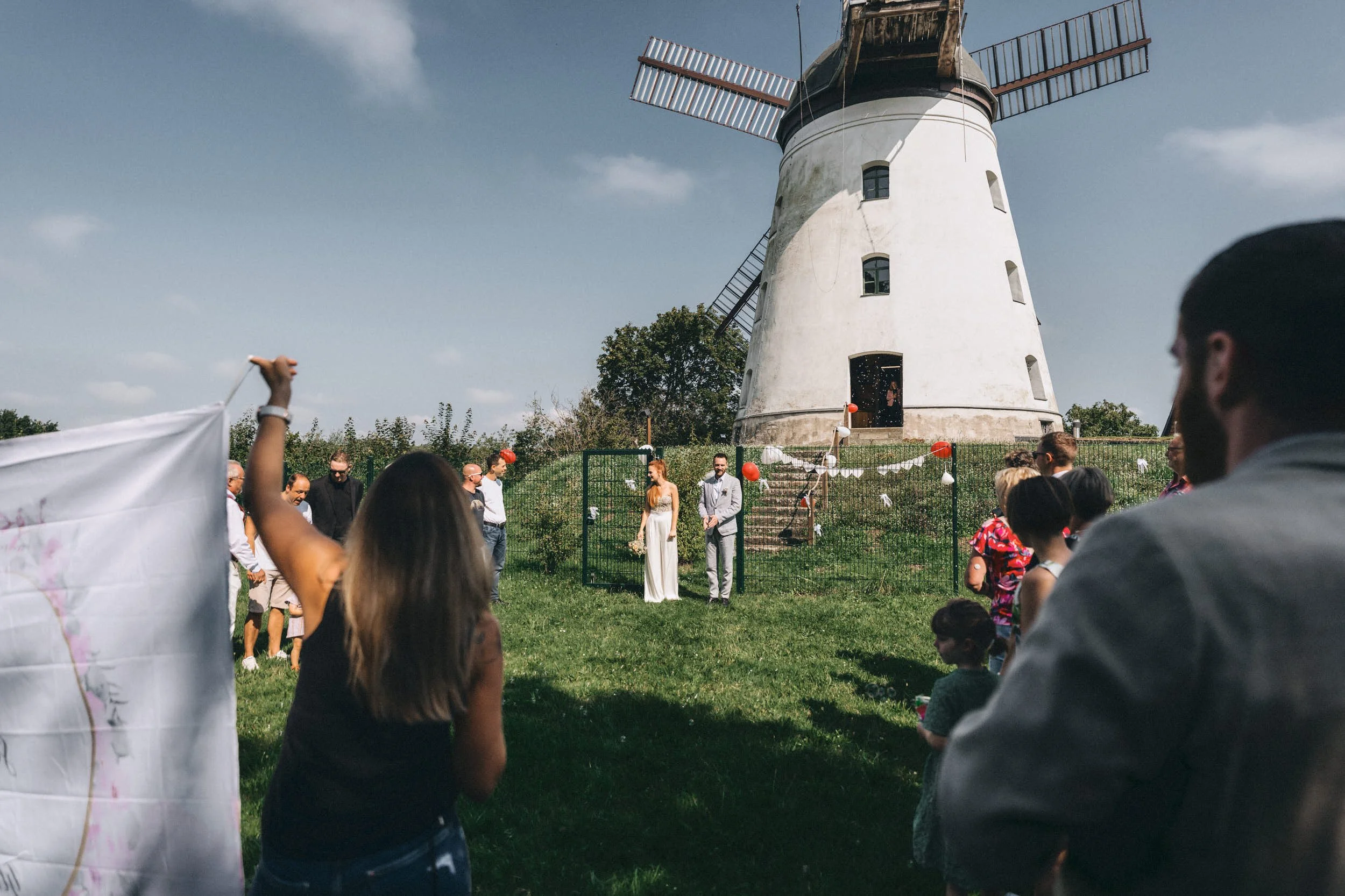 Ein Brautpaar bei einer Hochzeit im Freien vor einer Windmühle, umgeben von Gästen.
