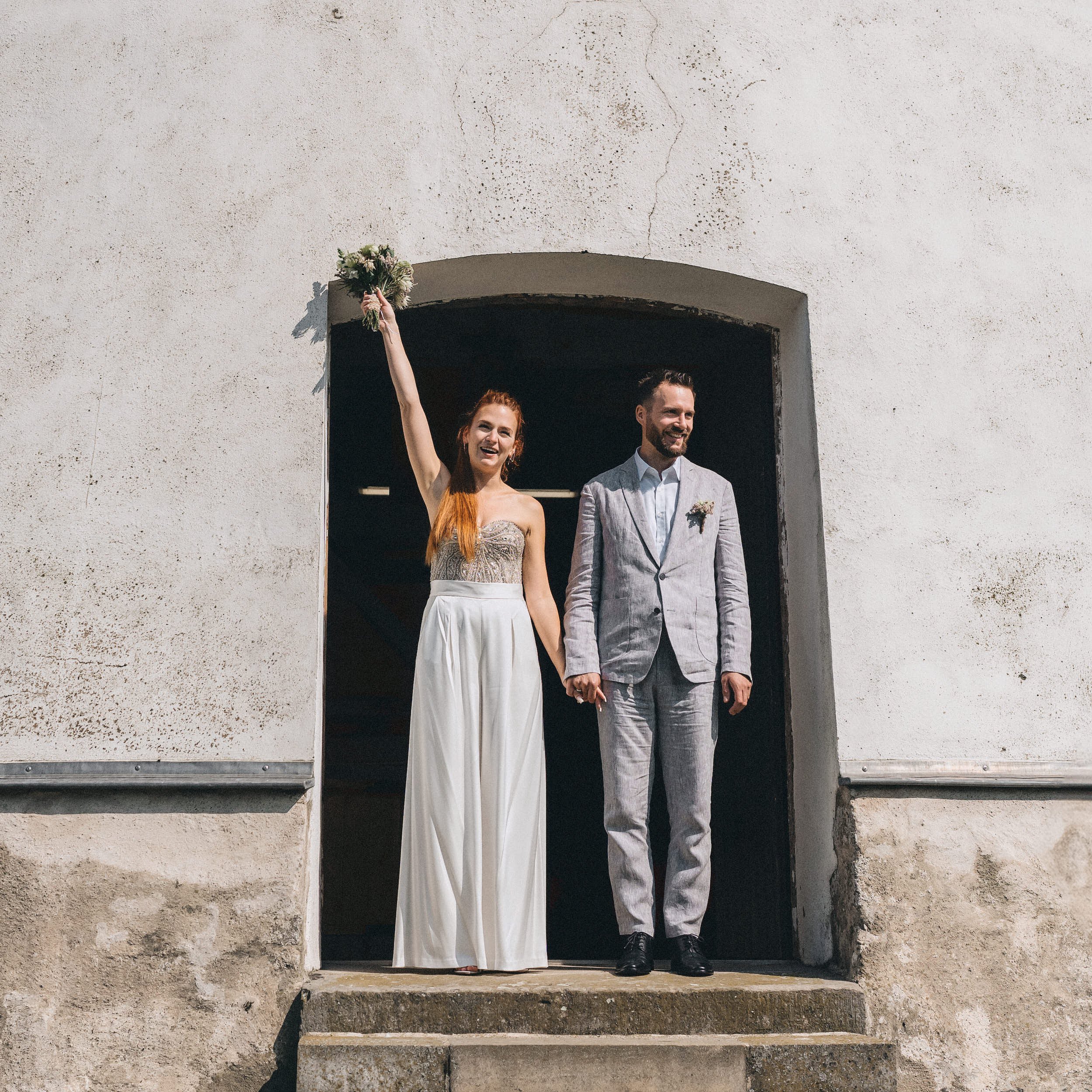 Ein Hochzeitspaar steht unter einem Bogen, die Braut hebt den Arm mit einem Blumenstrauß hoch, während der Bräutigam neben ihr steht. Sie sind vor einer weißen, leicht strukturierten Wand.