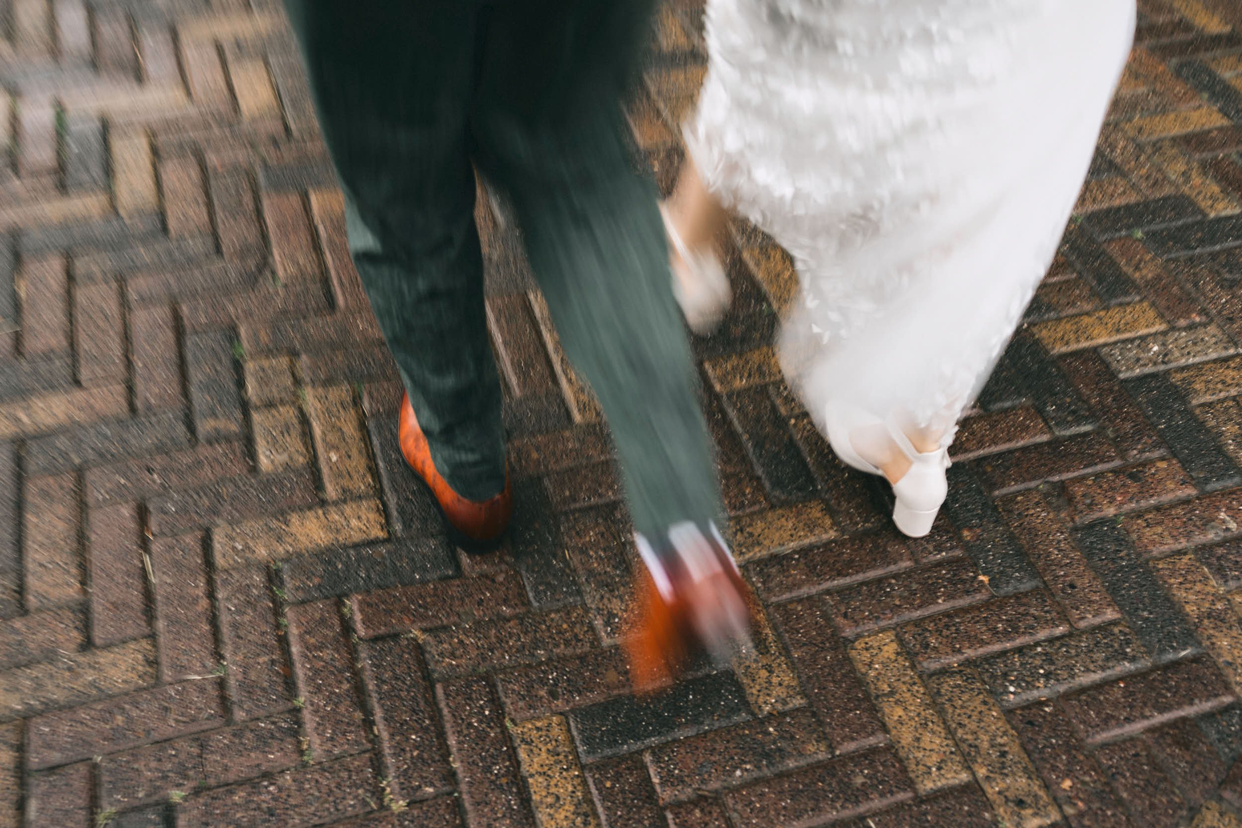 Menschen gehen über nassen, bunten Ziegelsteinpflaster bei Regen, eine Person trägt rote Schuhe, eine andere trägt weiße Kleidung.