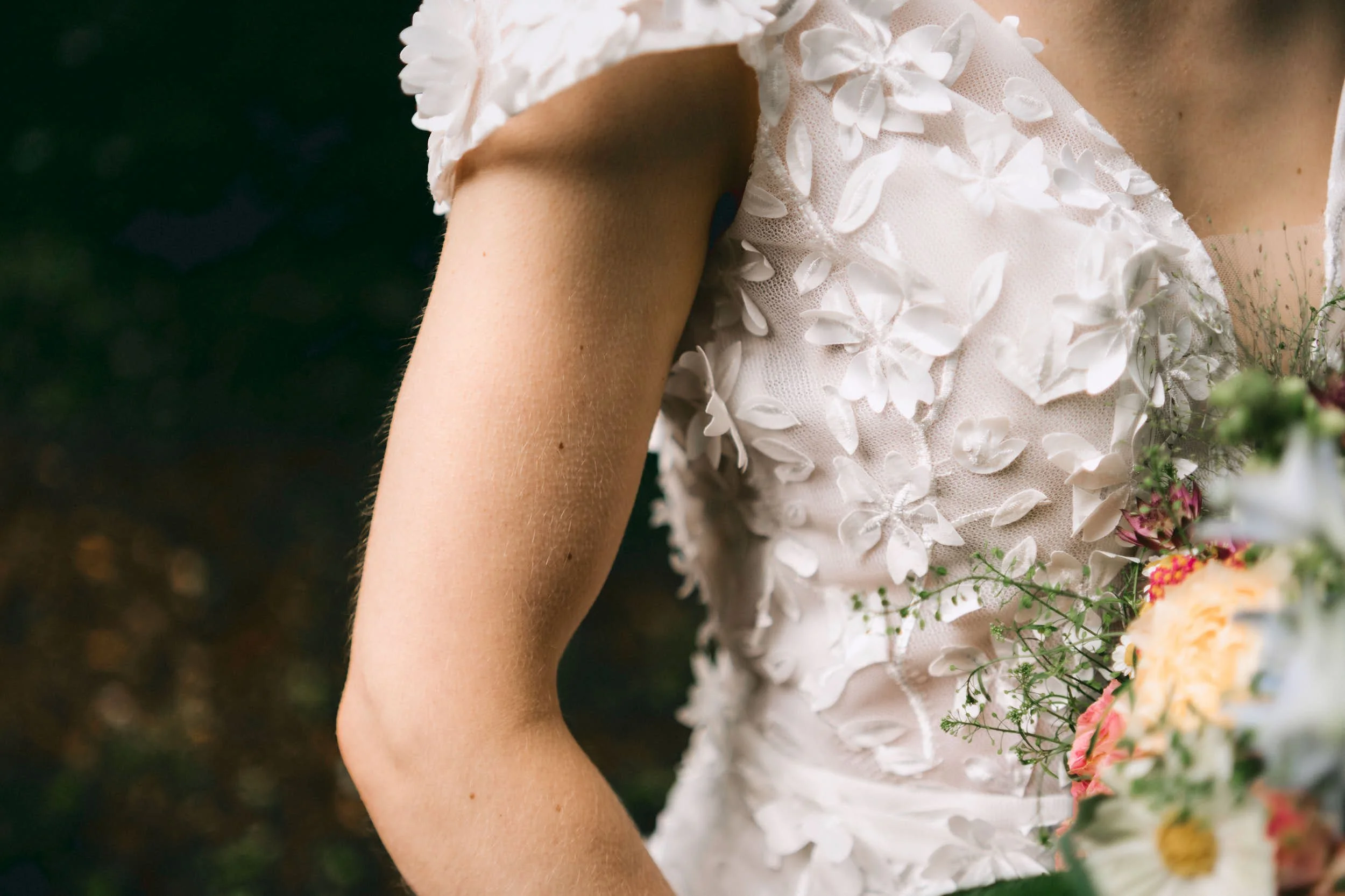 Nahaufnahme eines weißen Brautkleids mit 3D-Blumenapplikationen und eine Frau, die einen Blumenstrauß hält.