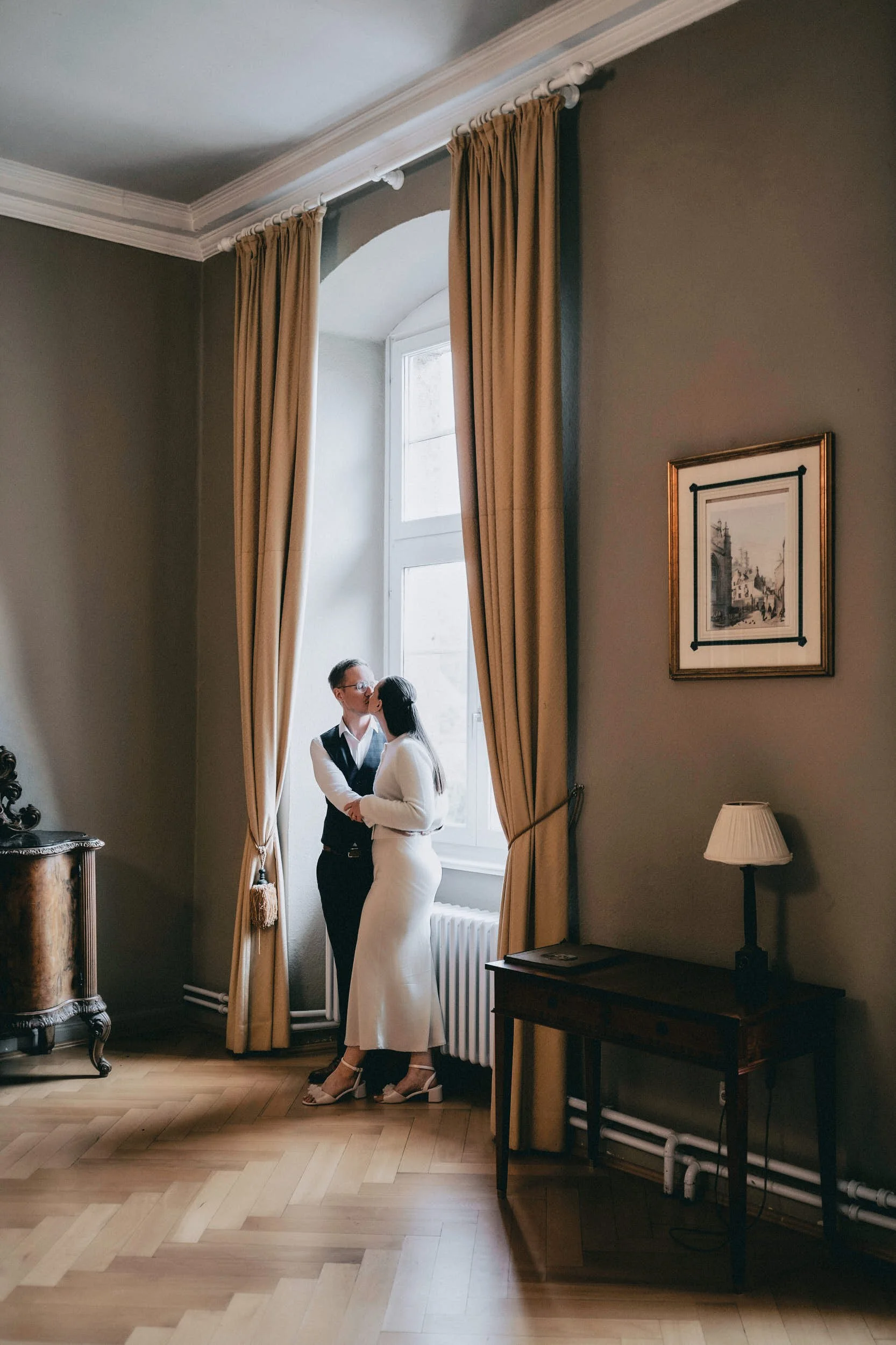 Ein Mann und eine Frau stehen sich gegenüber vor einem großen Fenster mit Sofortvorhängen. Sie scheinen sich zu küssen. Die Frau trägt ein weißes Hochzeitskleid, der Mann trägt dunkle Hemd und Weste. Der Raum hat hellen Holzboden, ein Wandbild und ei